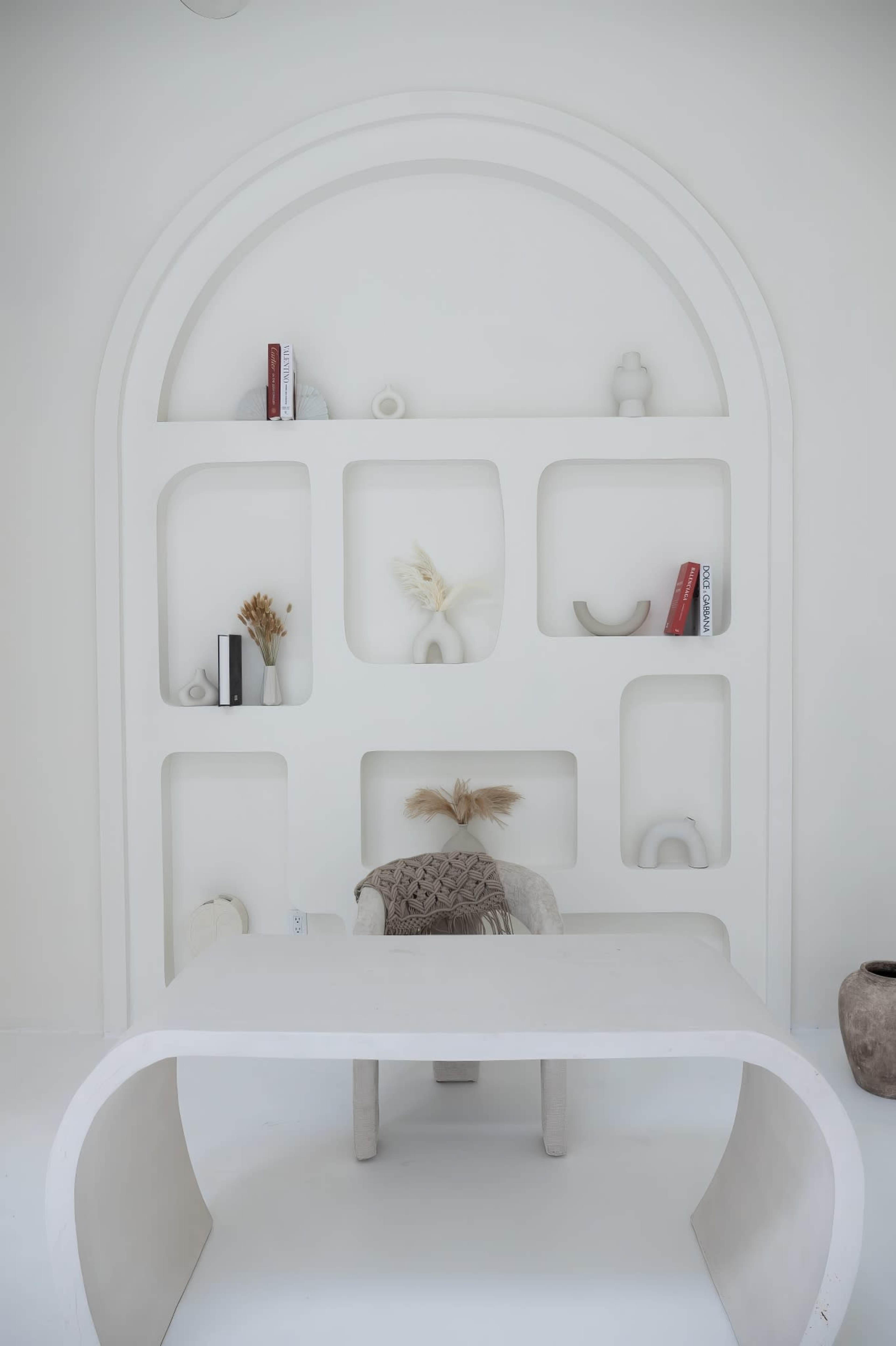 A minimalist workspace features a white desk in front of a curved shelving unit displaying decorative items and books.