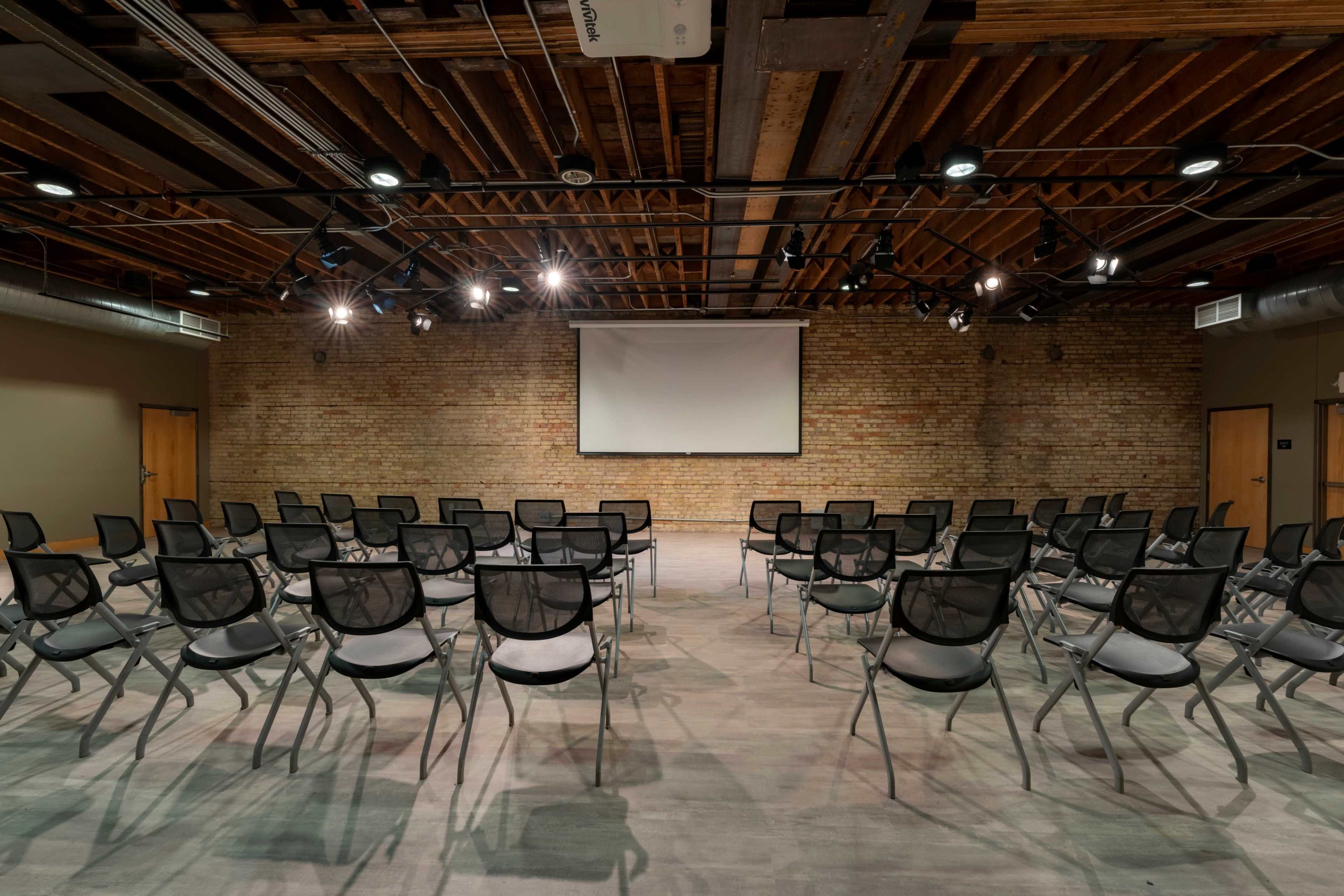 A meeting room with rows of black mesh chairs facing a projector screen on a brick wall.