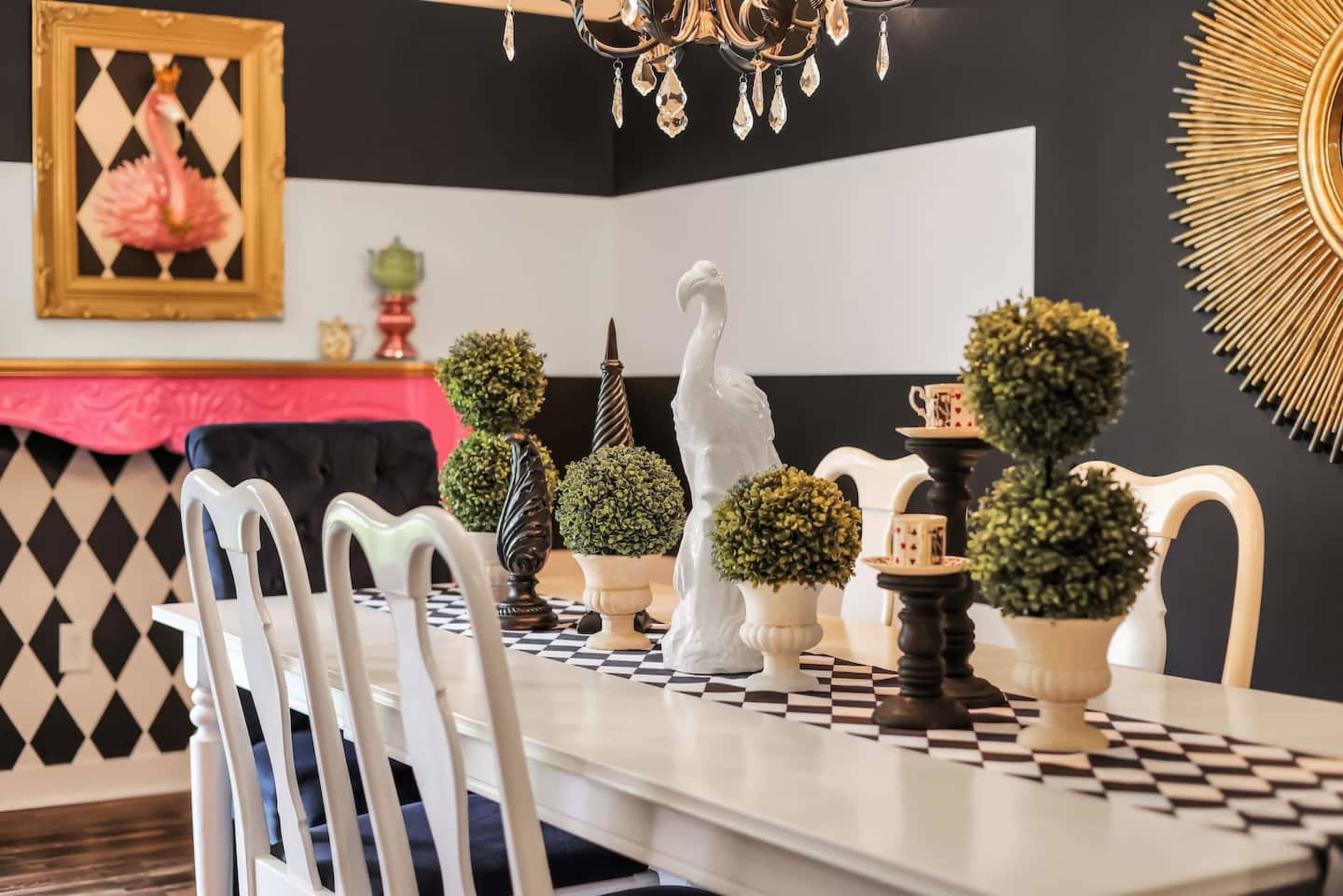A dining table is set with topiary centerpieces, decorative sculptures, and a black and white checkered runner under a chandelier in a room with dark walls and colorful accents.