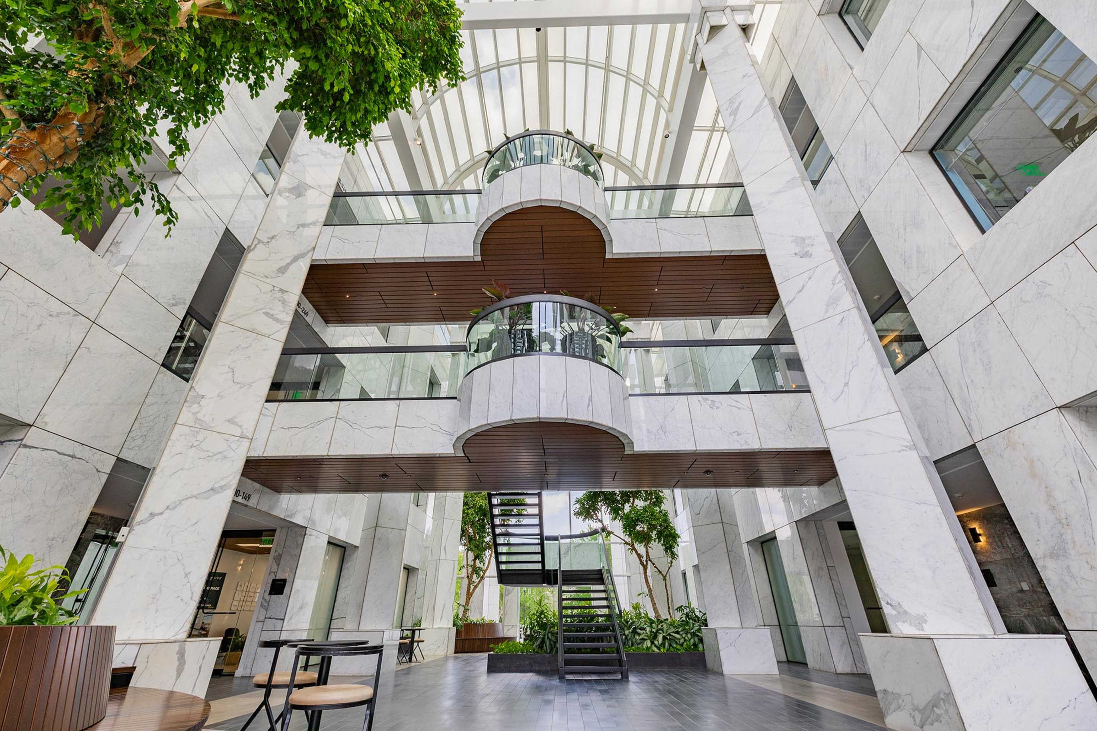 The image shows a modern interior space featuring marble walls, multiple levels with glass railings, and a central staircase surrounded by greenery.