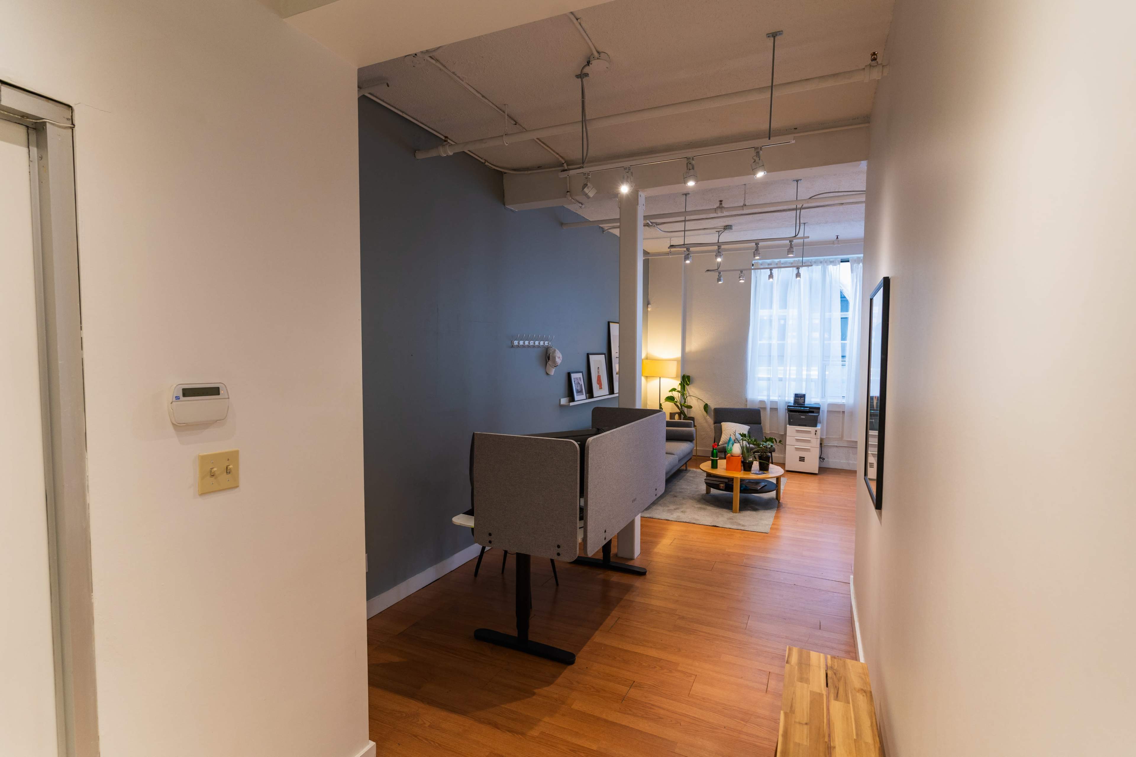 The image shows a hallway leading to a modern living room with a gray accent wall, a sofa, a coffee table, and large windows allowing natural light to enter.