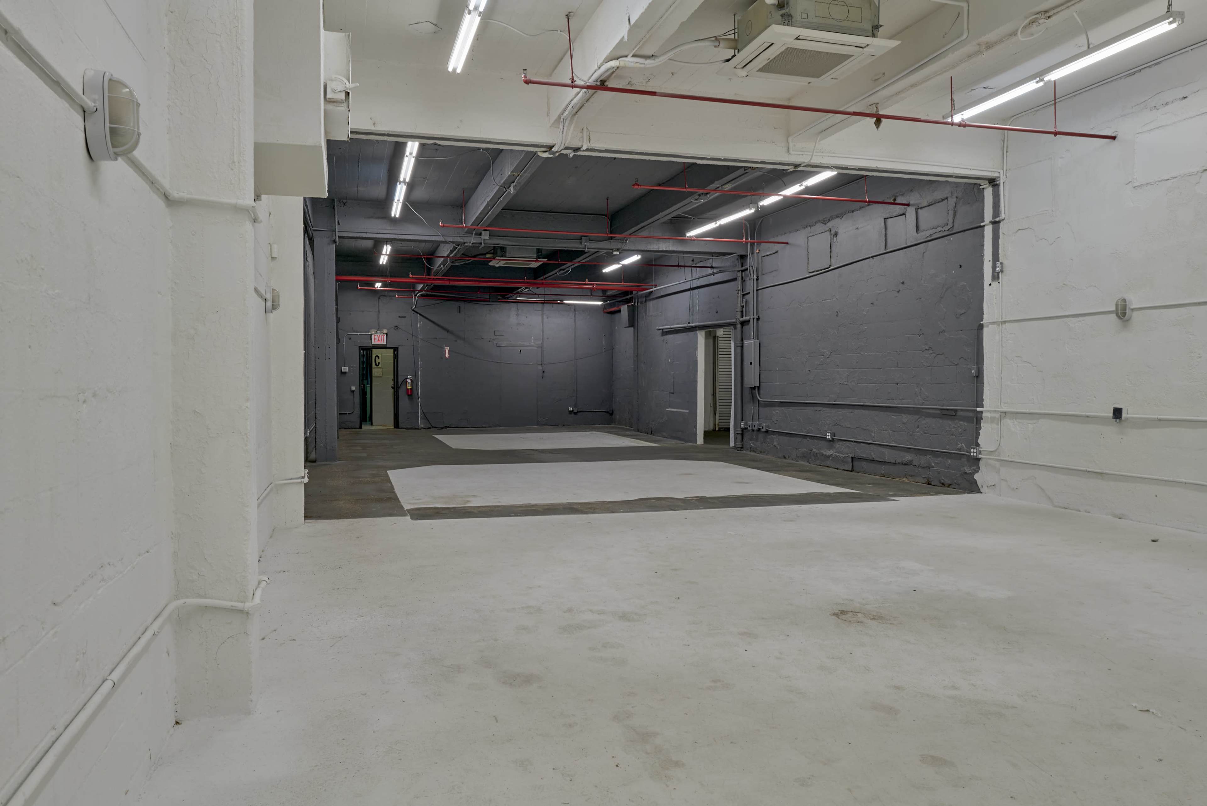 The image shows a large, empty industrial space with concrete floors and gray walls, illuminated by overhead fluorescent lights.