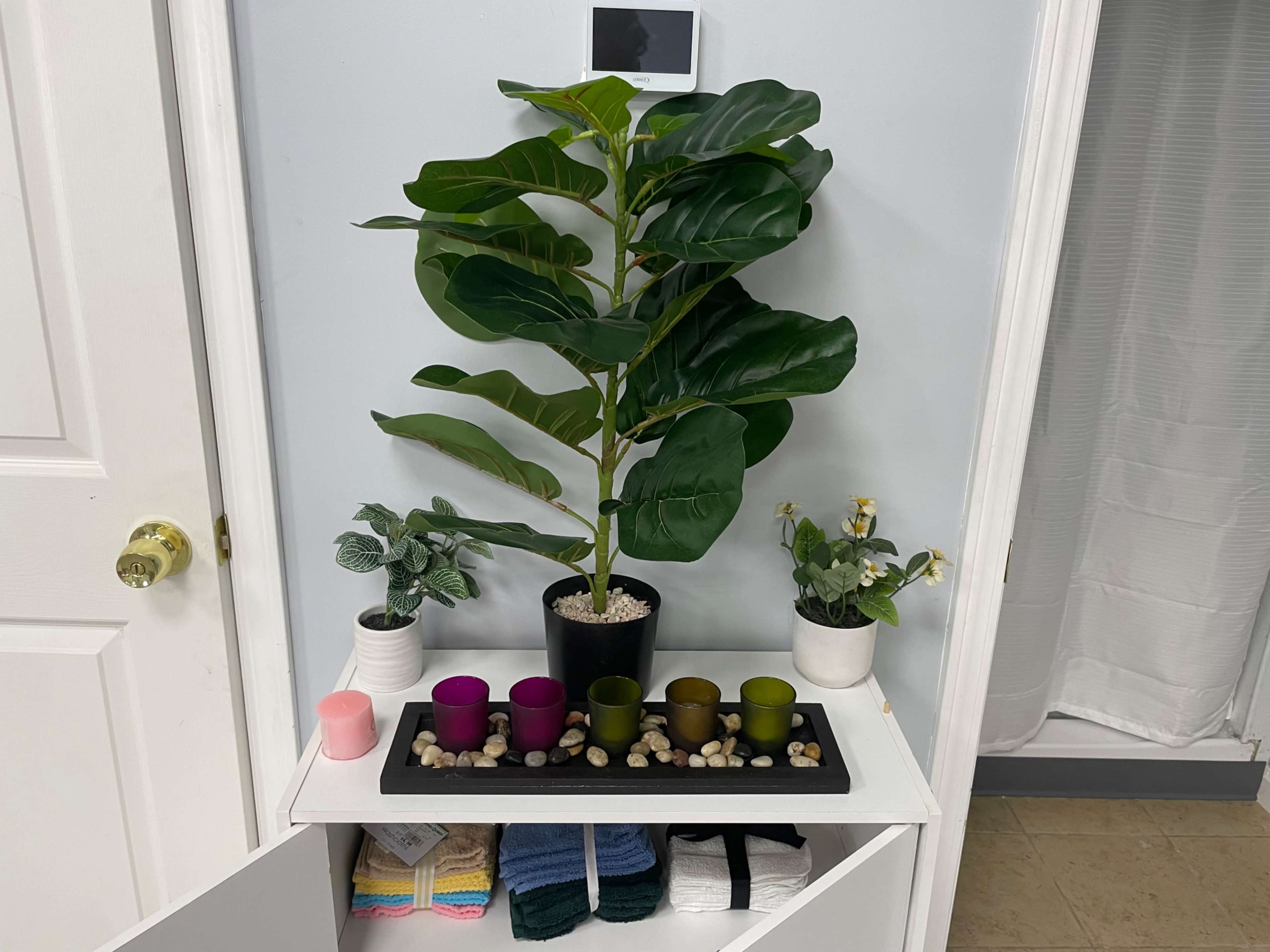 A decorative shelf features a large artificial plant, several small potted plants, colorful candles, and neatly folded towels.