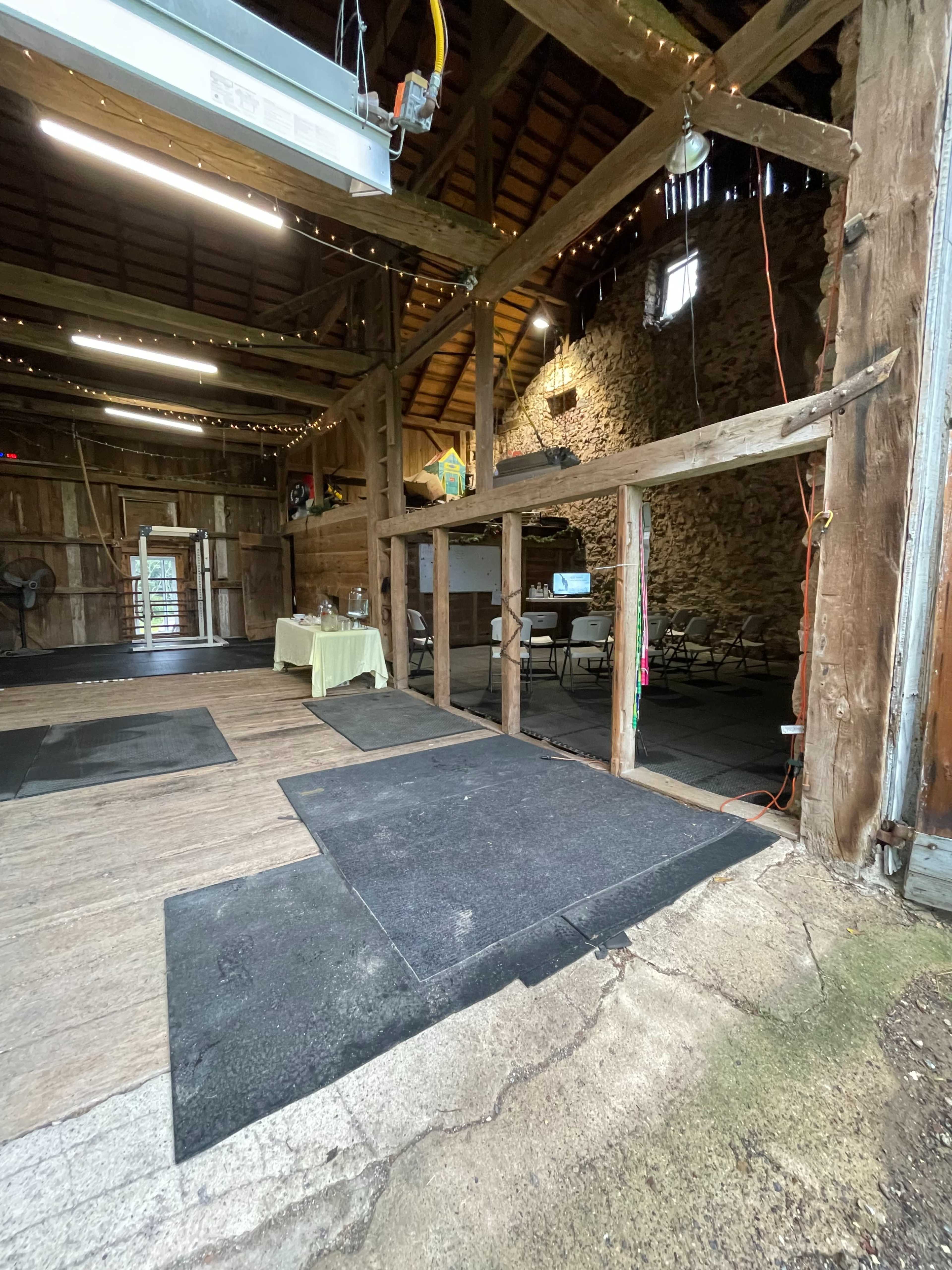 The interior of a wooden barn features gym mats on the floor and a table set up in one corner, with additional chairs visible in the background.