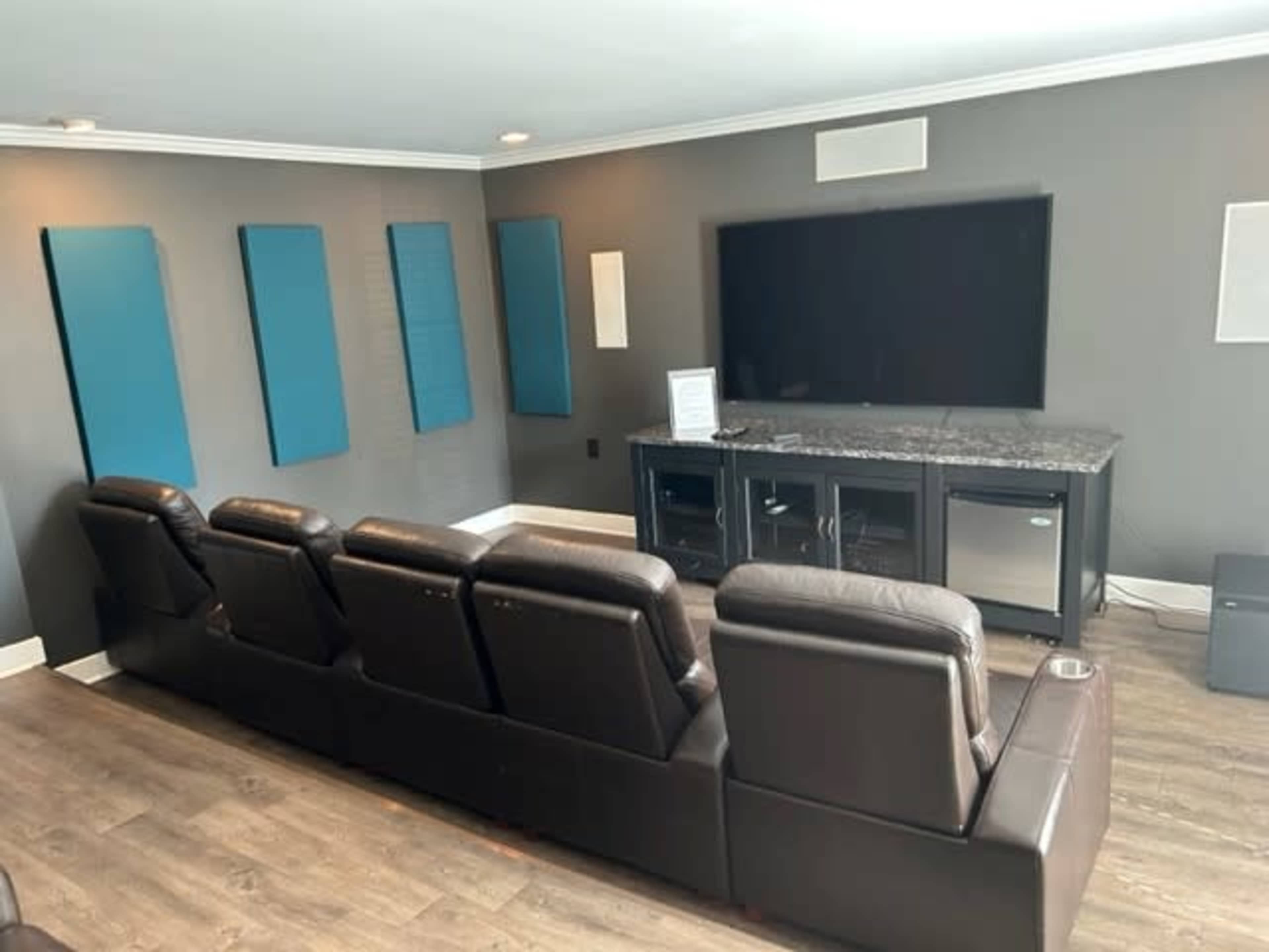 A modern media room with a row of leather recliners facing a large television mounted on a gray wall, complemented by soundproofing panels.