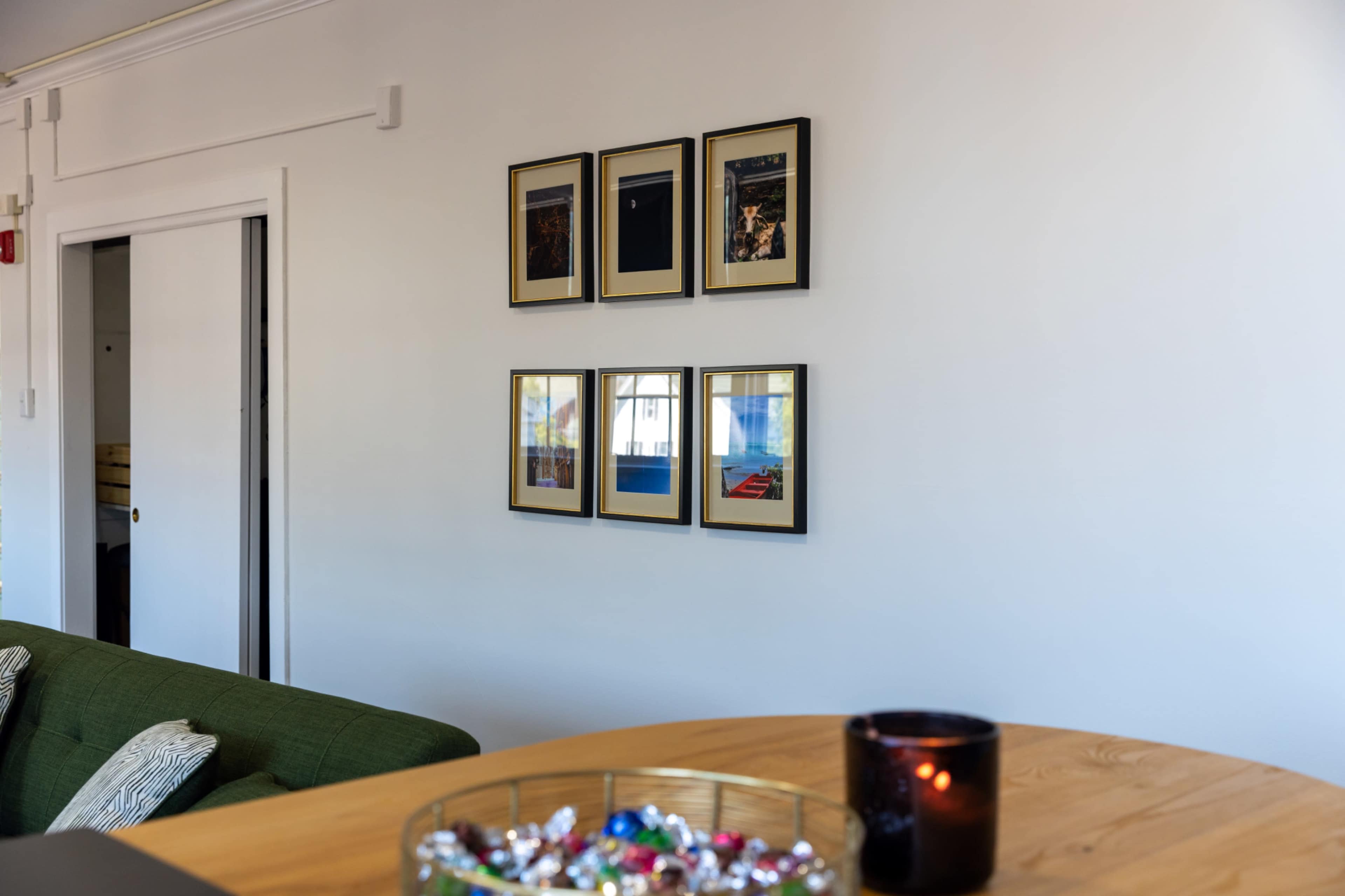 The image shows a wall with six framed pictures arranged in two columns, a green couch in the foreground, and a wooden table with a decorative bowl and a candle.