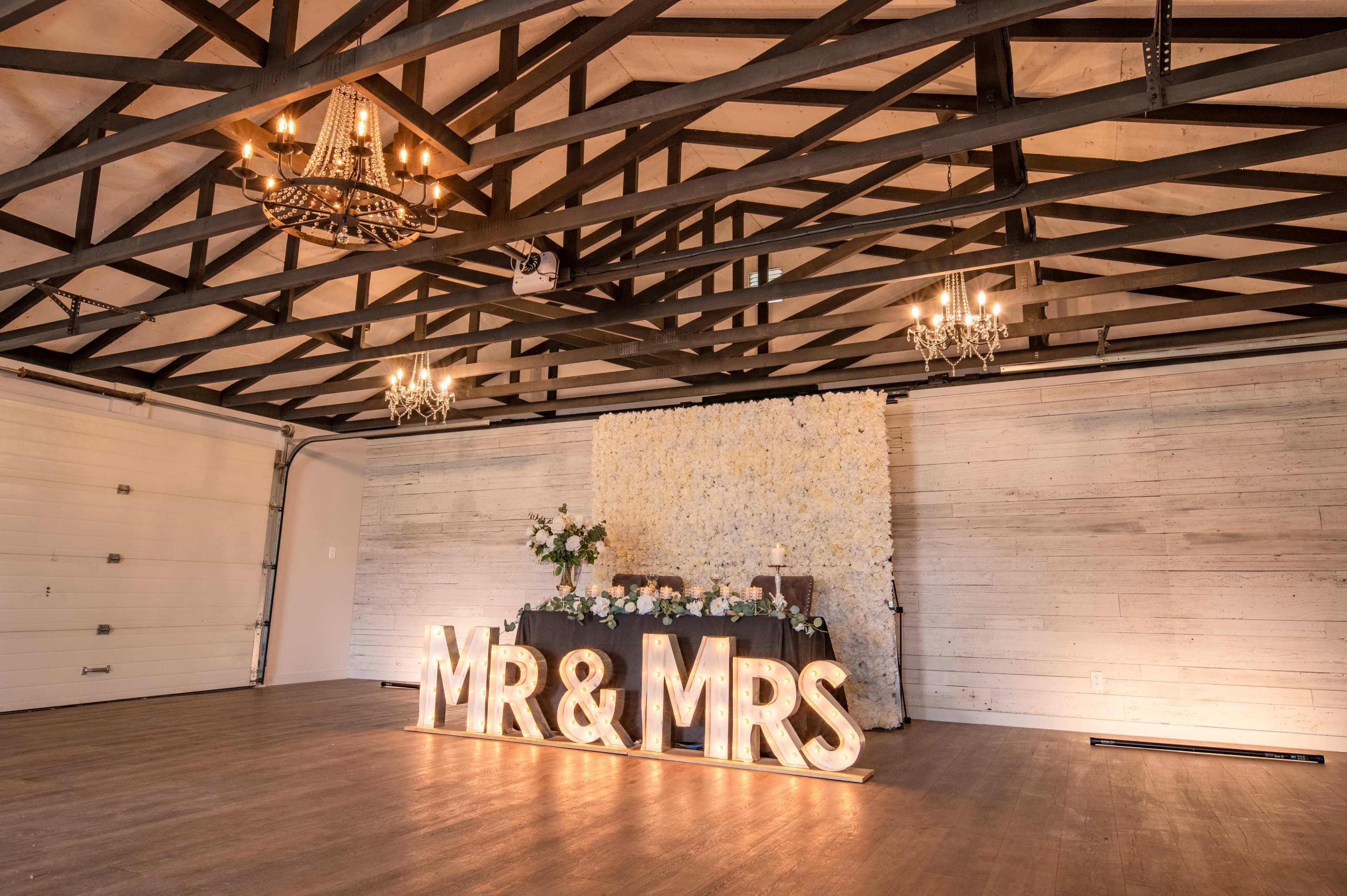 The image shows a wedding reception area featuring a decorative "MR & MRS" sign, a floral backdrop, and chandeliers hanging from a wooden-beamed ceiling.