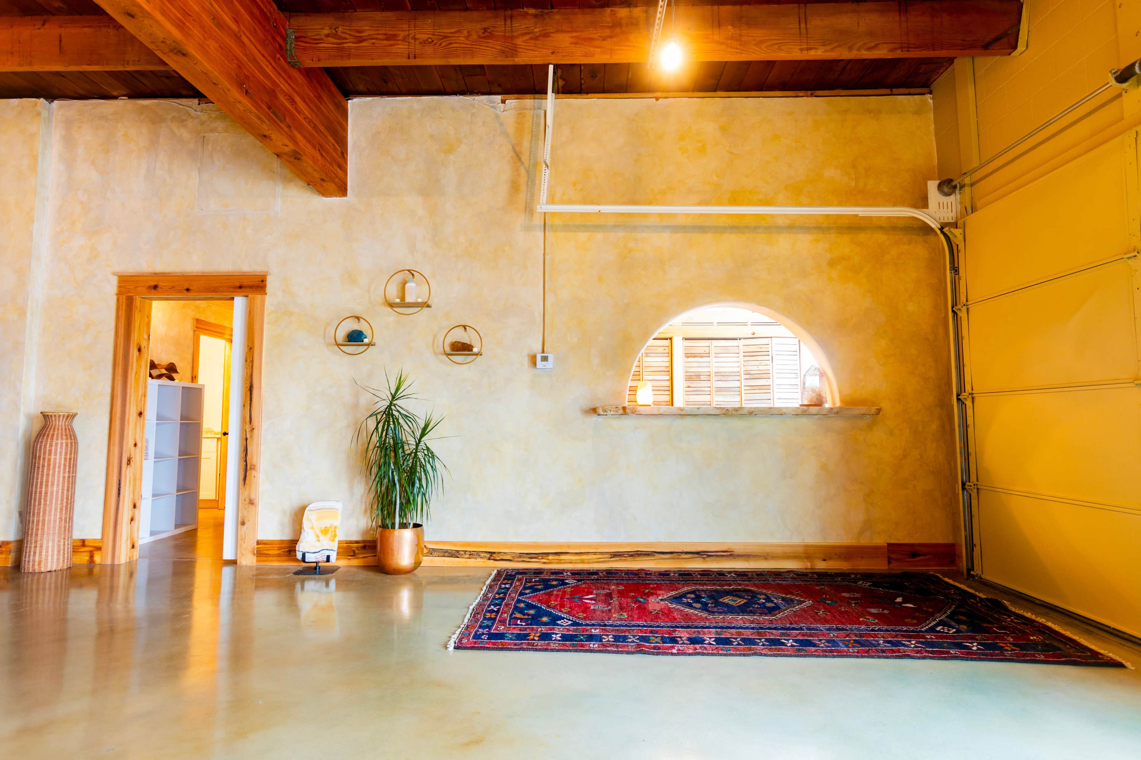 A spacious room with a concrete floor, light yellow walls, wooden beams, a decorative rug, and a plant next to an arched opening leading to another area.