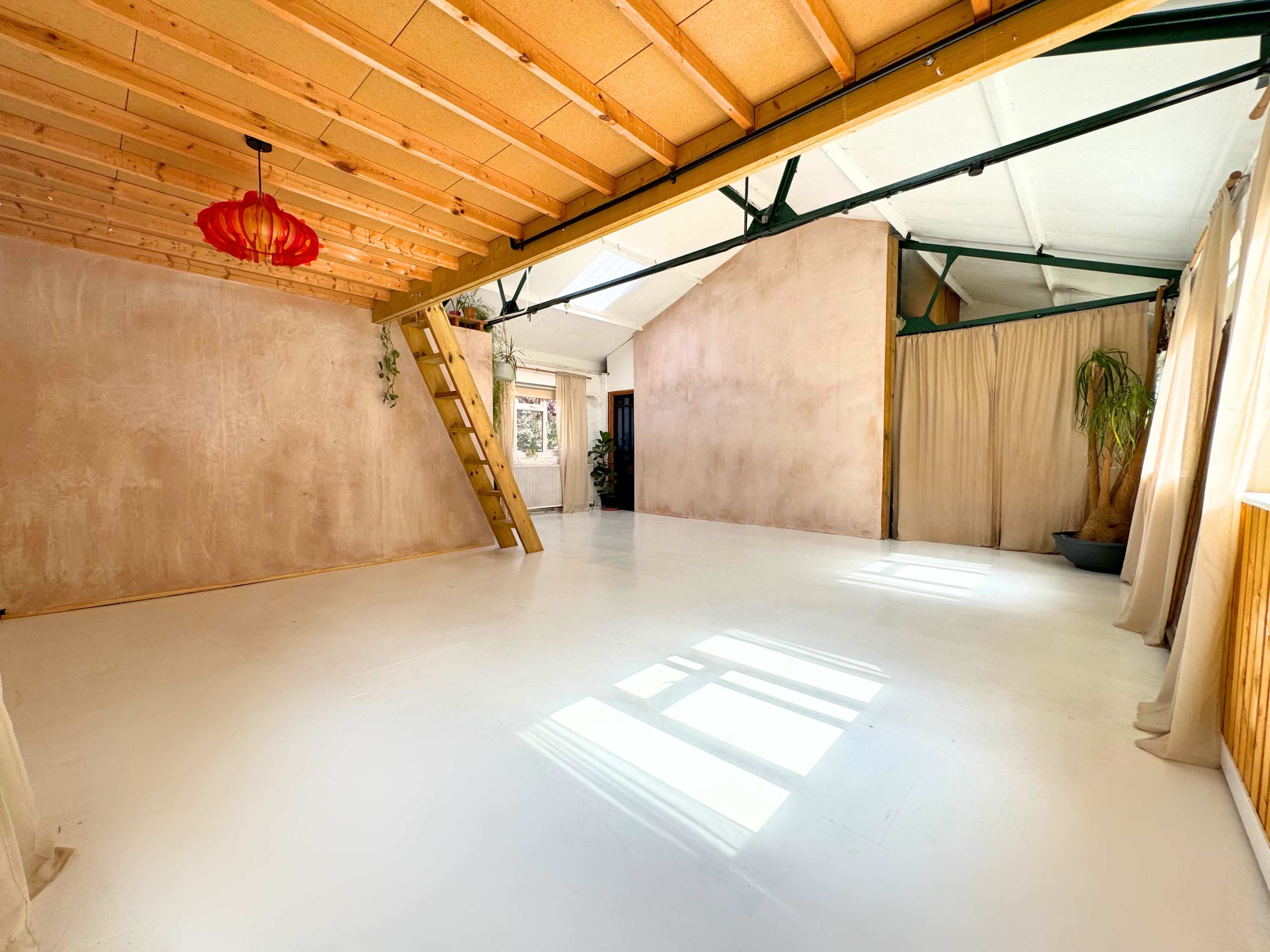 A spacious, sunlit room with wooden beams, a ladder leading to a loft, and large windows allowing natural light to fill the white-painted floor.