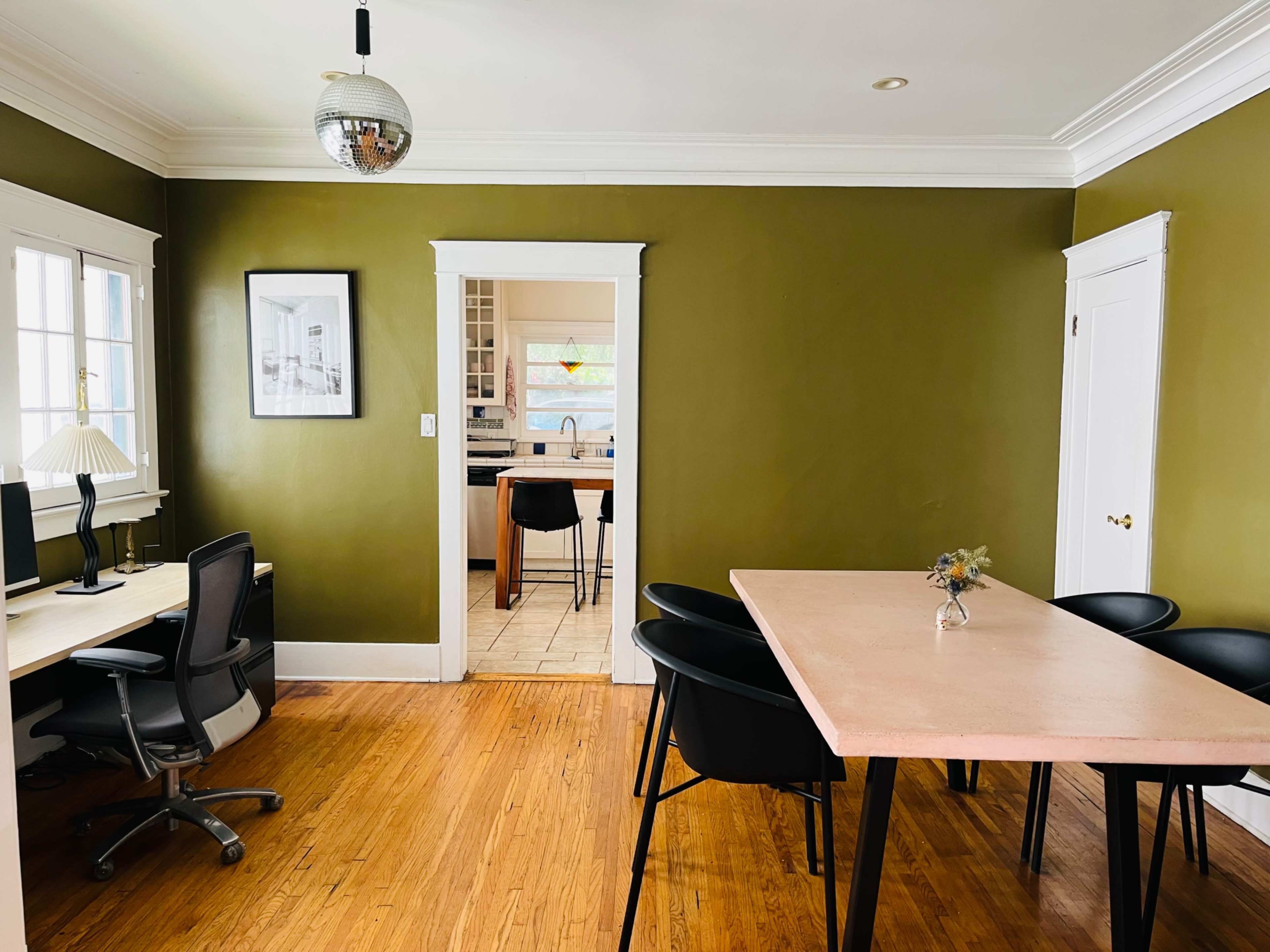 A room with a green wall, a wooden floor, a table surrounded by black chairs, and a doorway leading to a kitchen area.
