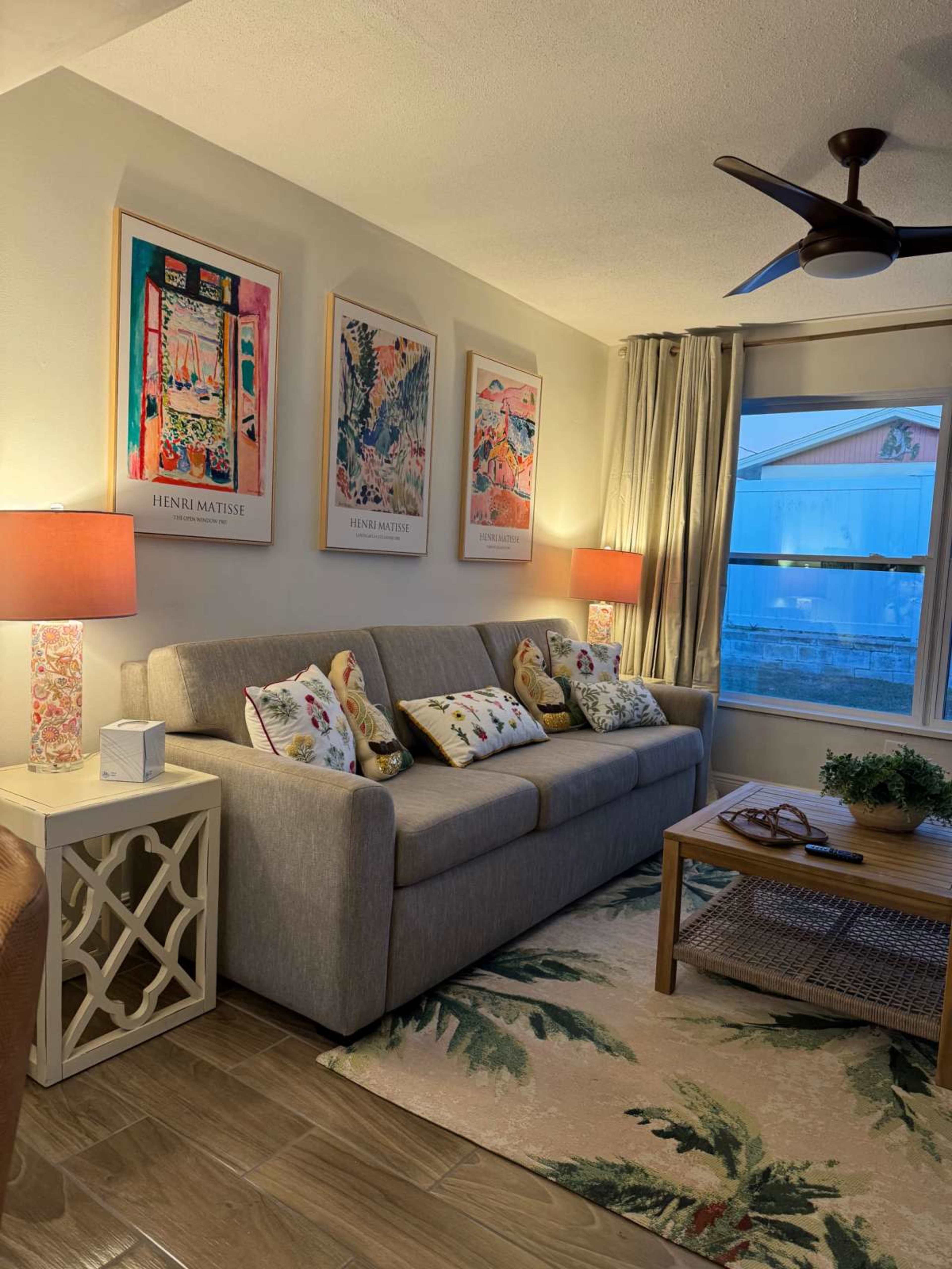 The image shows a living room with a light gray sofa adorned with decorative pillows, vibrant artwork on the walls, and a wooden coffee table on a patterned rug.