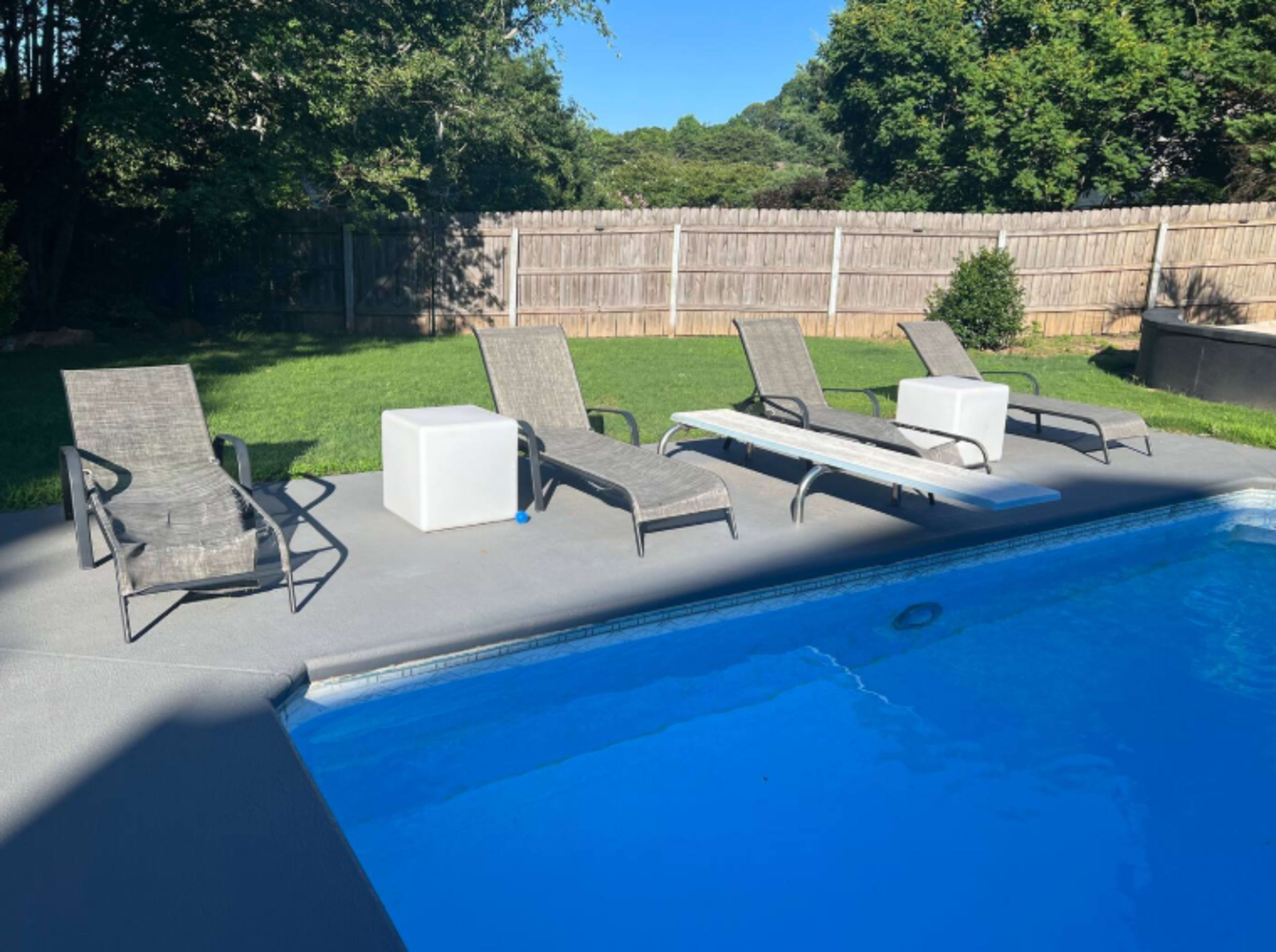 A clear blue swimming pool is surrounded by several lounge chairs on a sunlit patio with a grassy area and a wooden fence in the background.