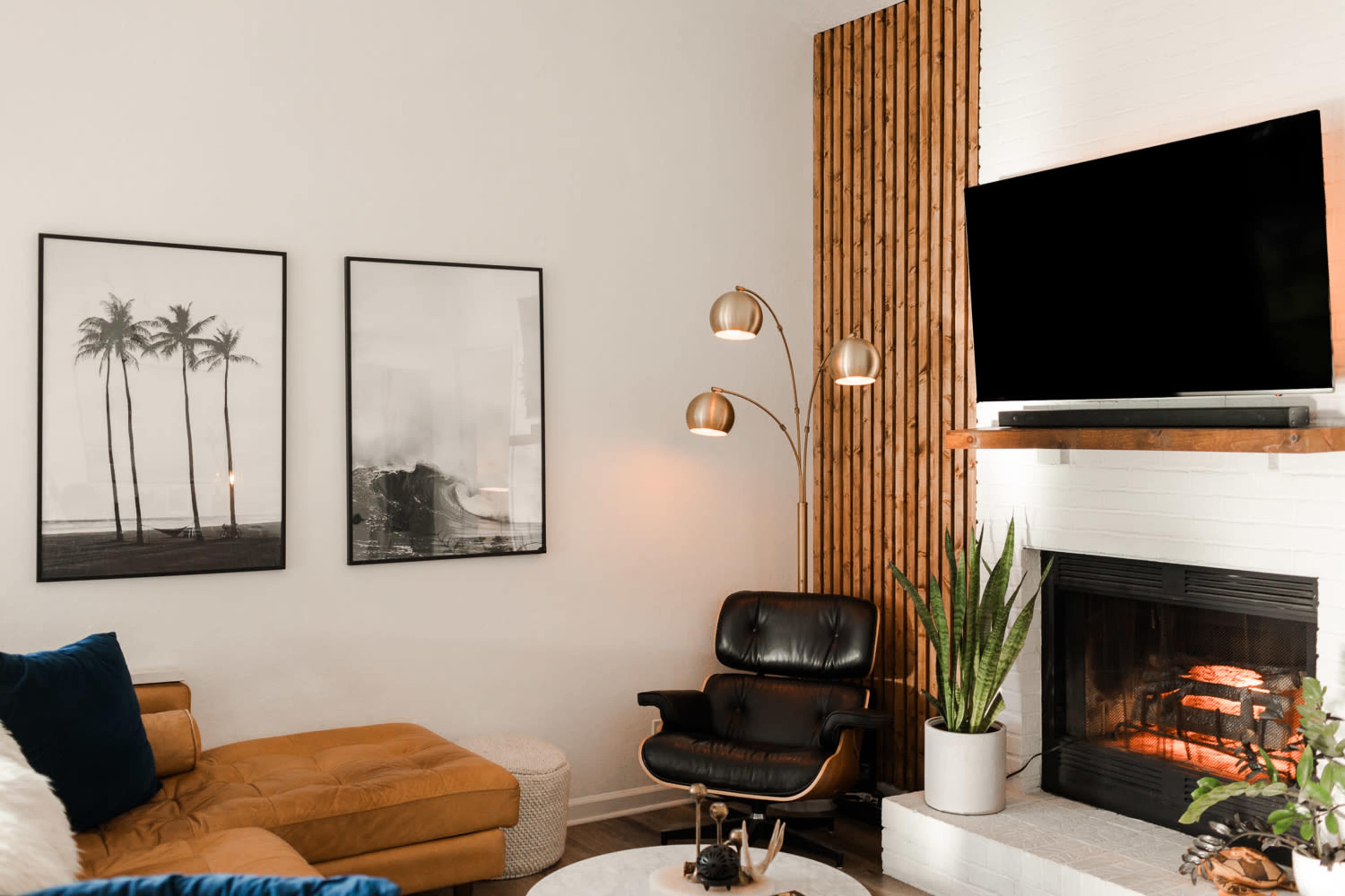 A modern living room features a tan sectional sofa, framed black-and-white palm tree photographs, a standing lamp, a television mounted above a wooden shelf, and a fireplace with a potted plant nearby.