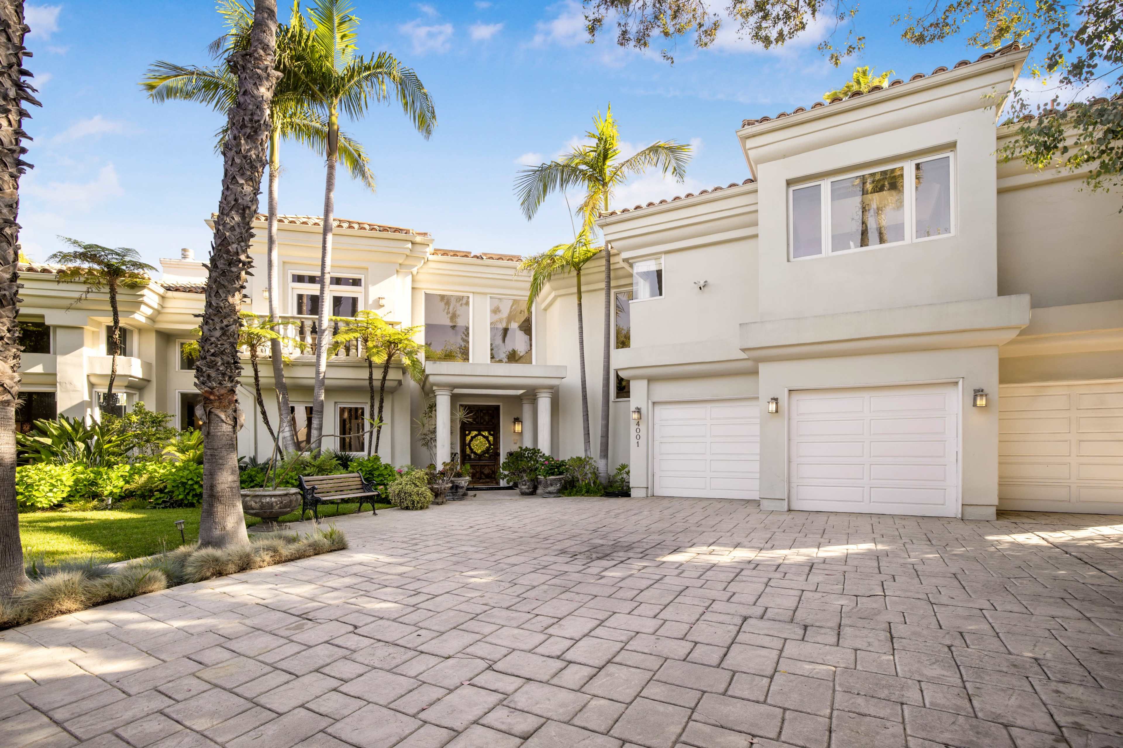 A modern two-story house with a paved driveway, palm trees, and a manicured garden.