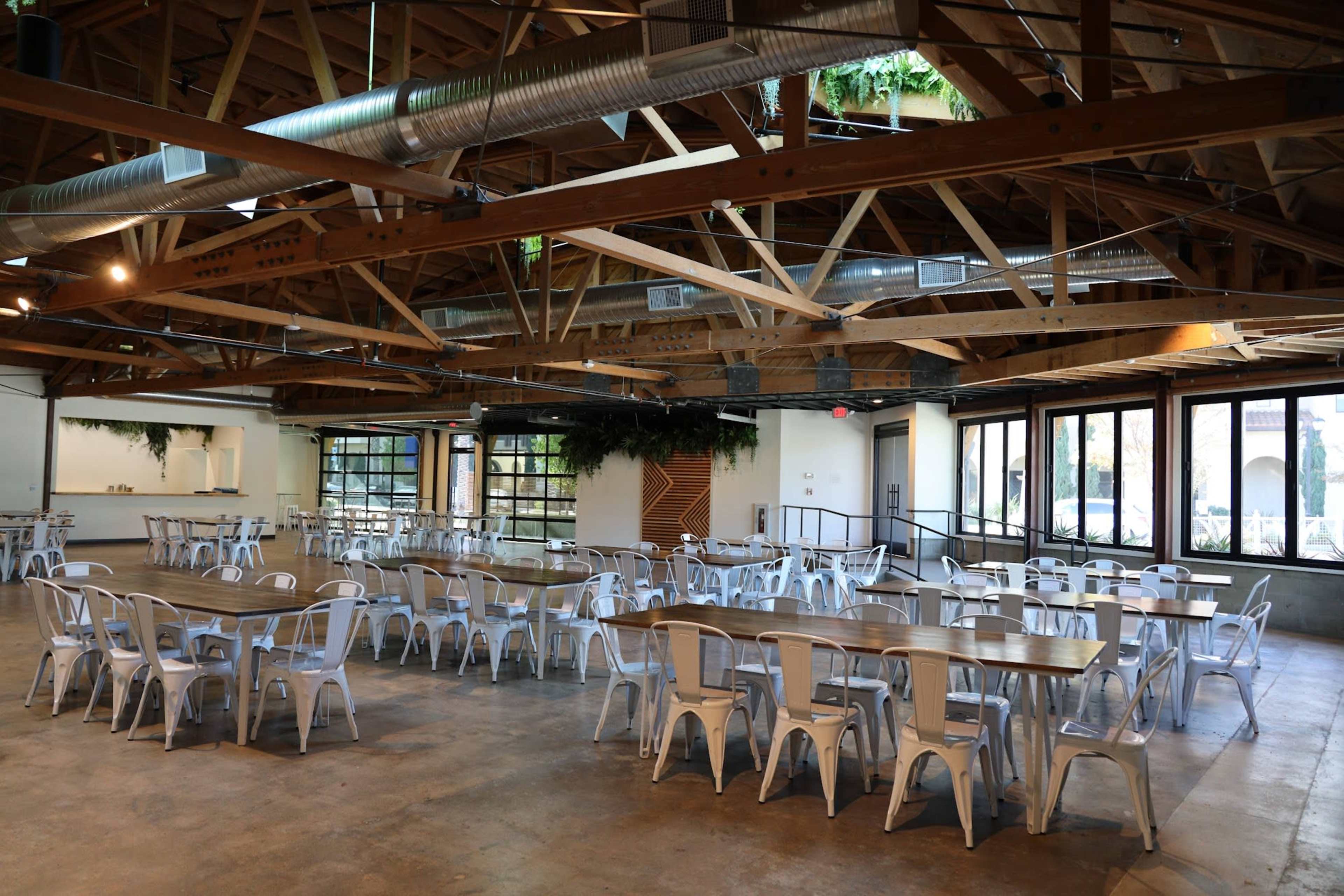 The image shows a spacious indoor event venue with multiple long tables and metal chairs arranged under a wooden beam ceiling.