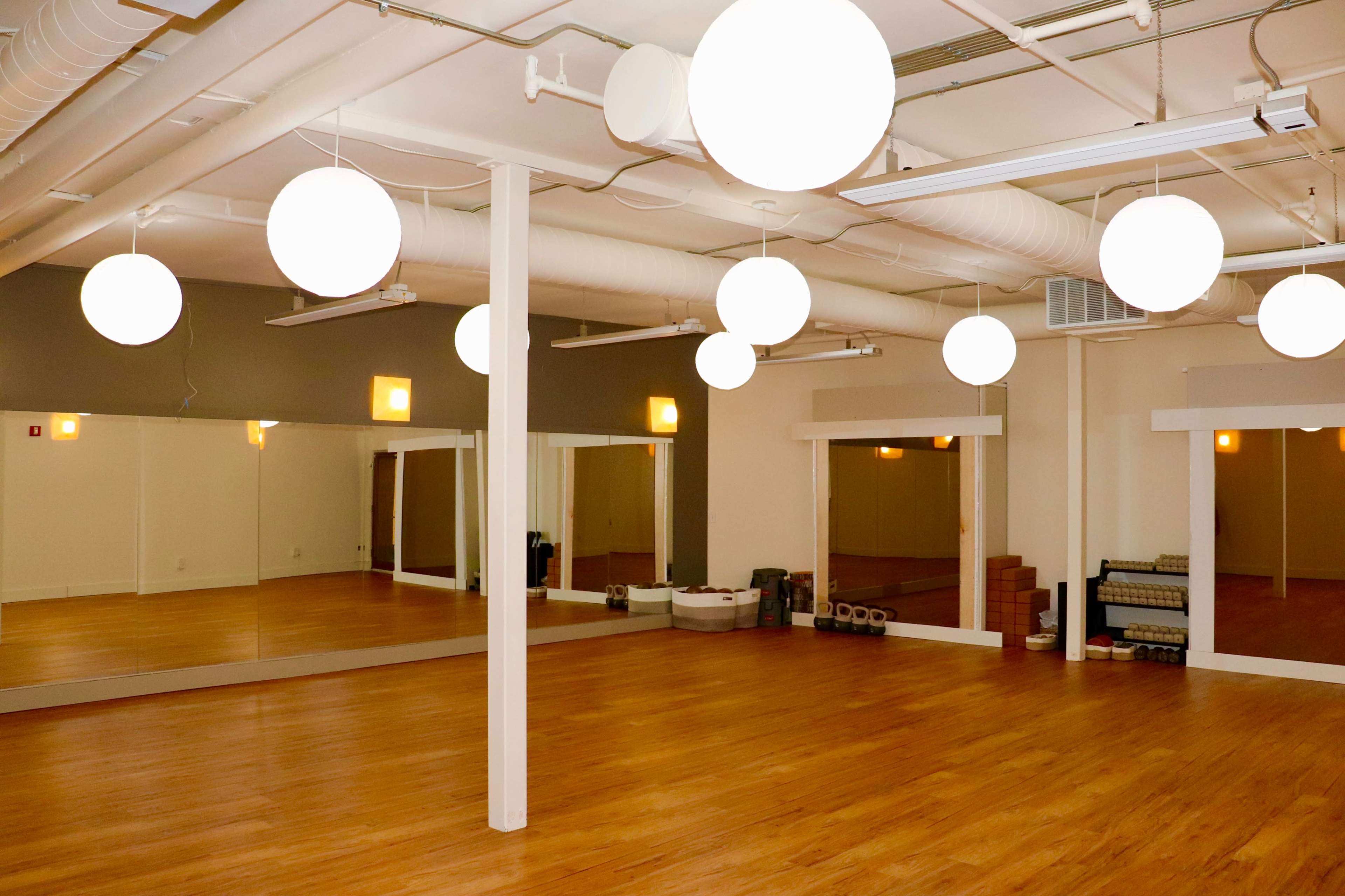 The image shows a spacious, well-lit studio with wooden flooring, large mirrors, and round white pendant lights hanging from the ceiling.