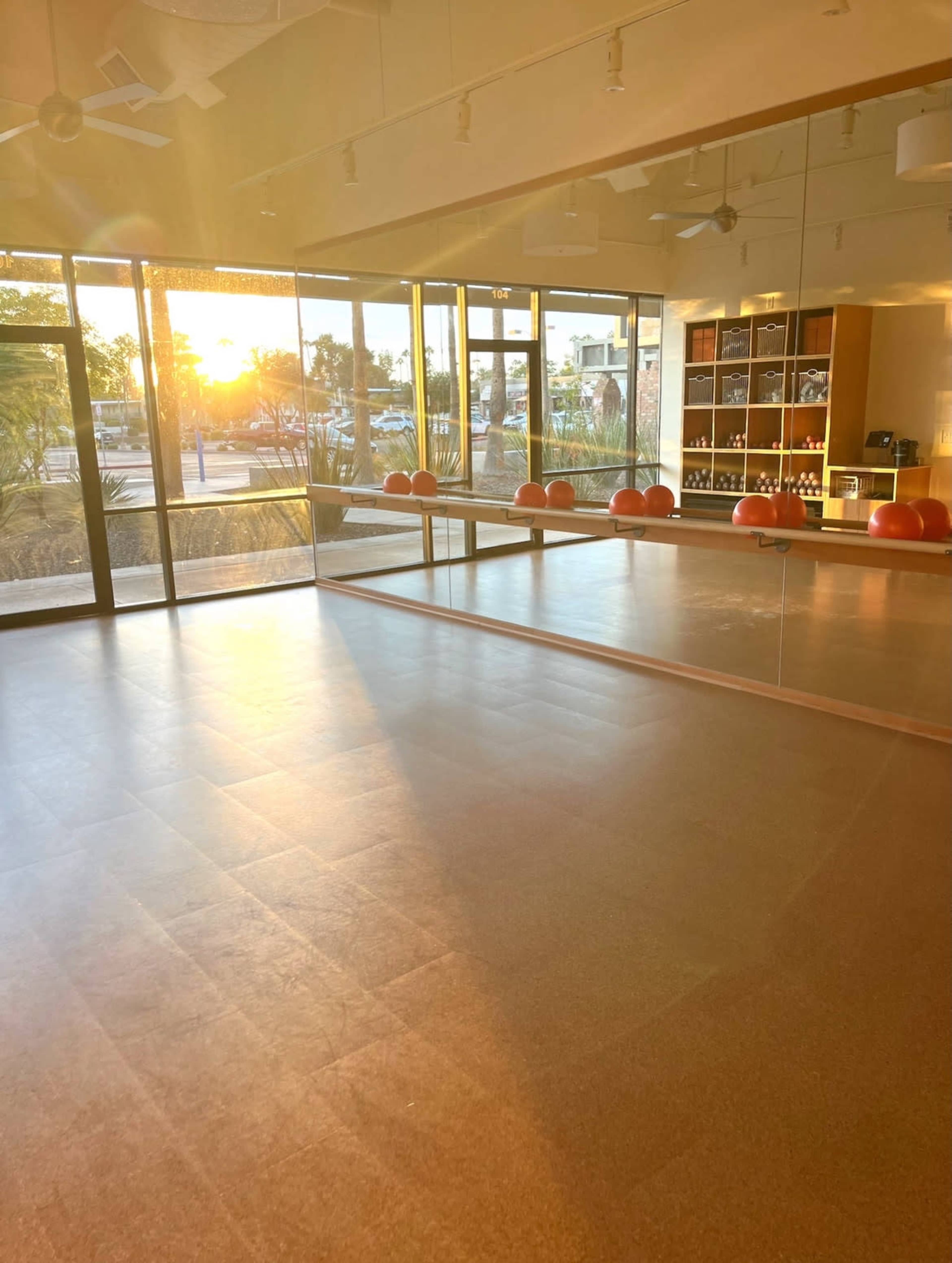 The image shows an empty exercise studio with large windows, reflecting a sunset and illuminated by warm golden light.