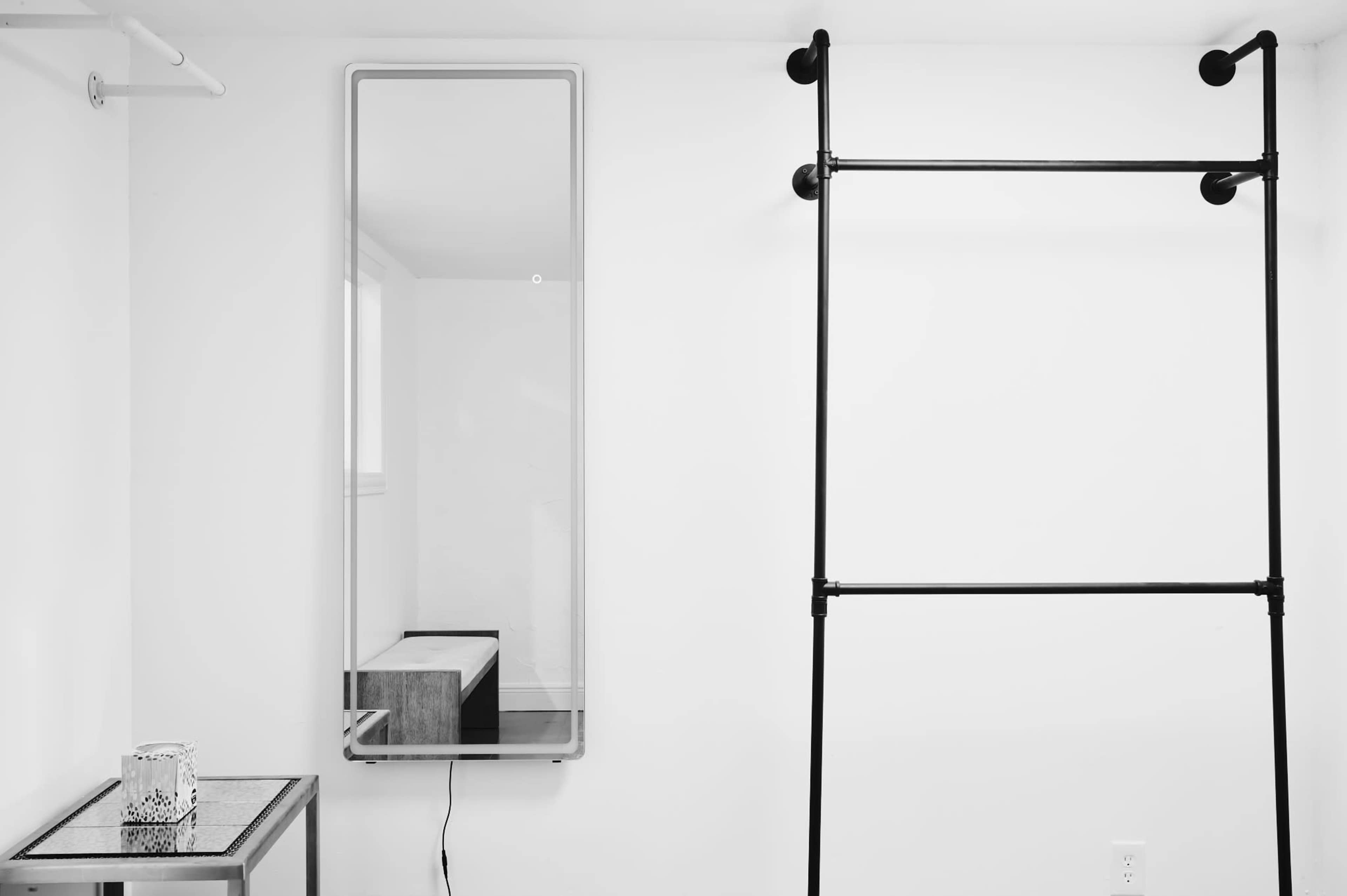 A minimalist interior featuring a tall, narrow mirror, a wooden bench, and a black clothing rack against a white wall.