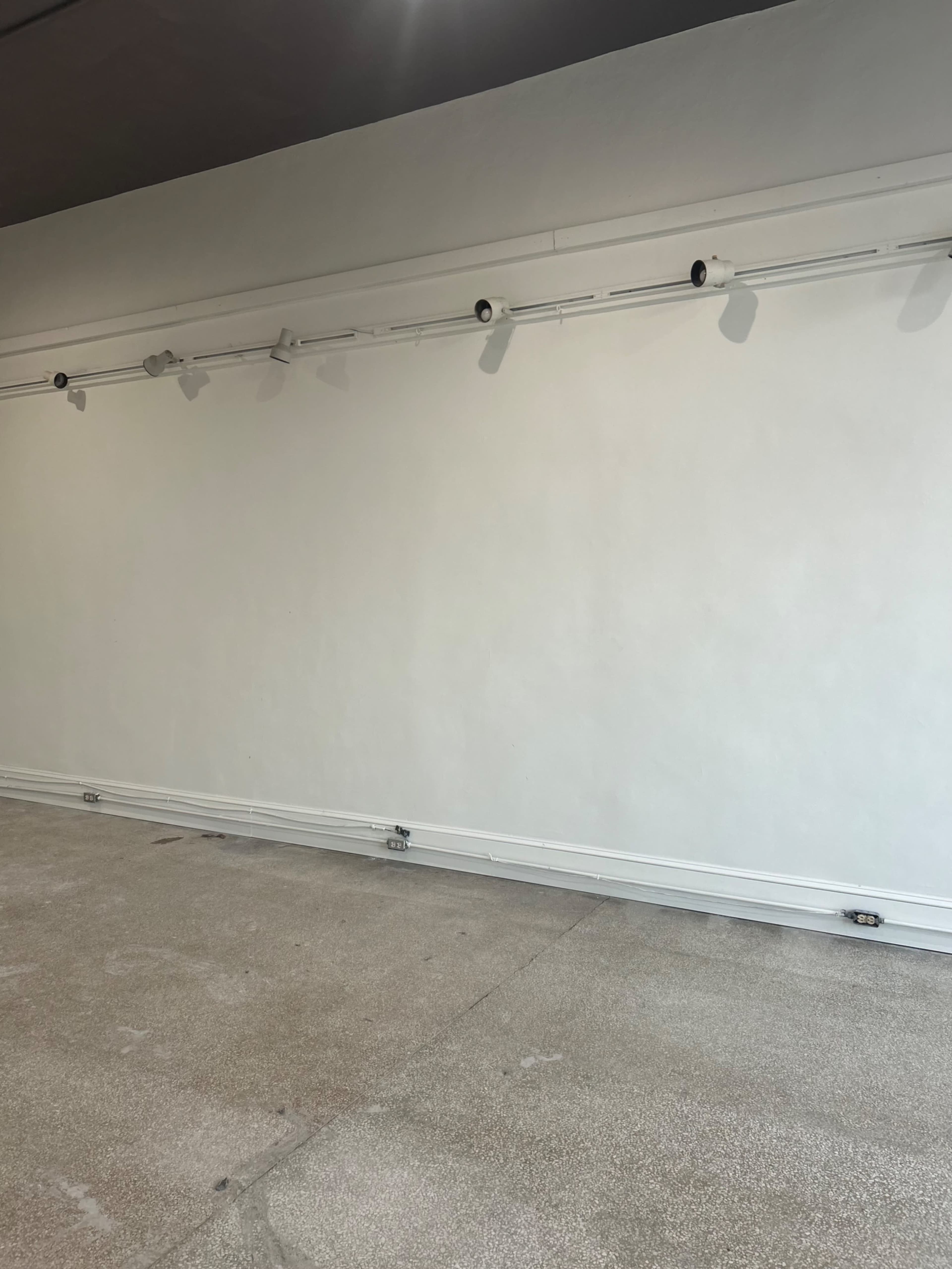 The image shows an empty interior wall with track lighting above and a bare concrete floor.