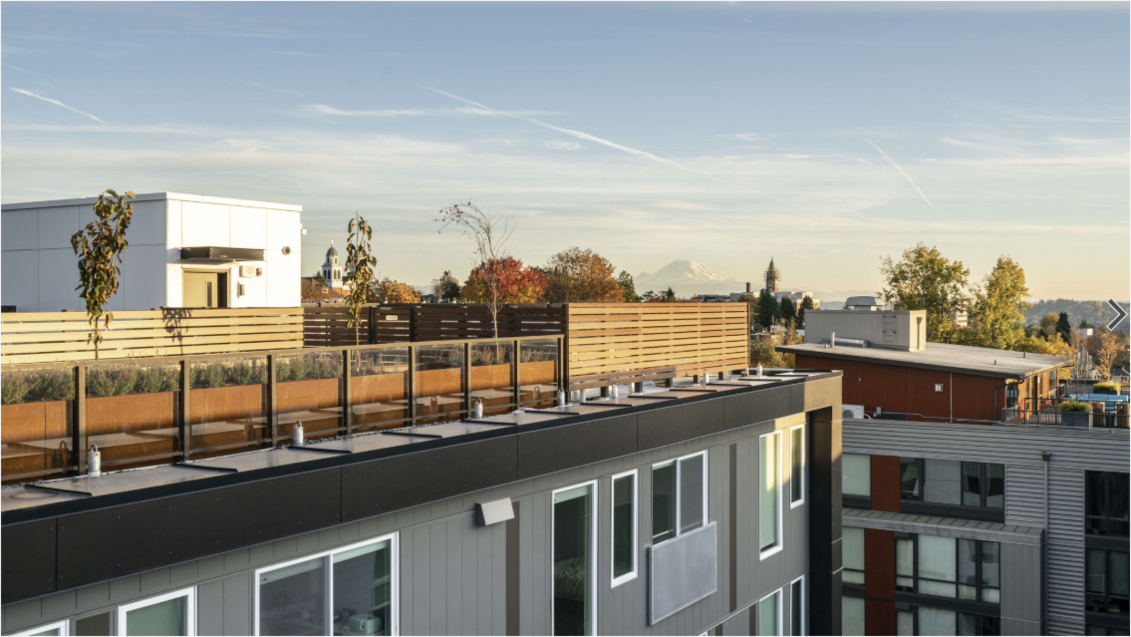 The image shows a modern building rooftop with wooden fencing and a view of distant mountains and trees in the background.