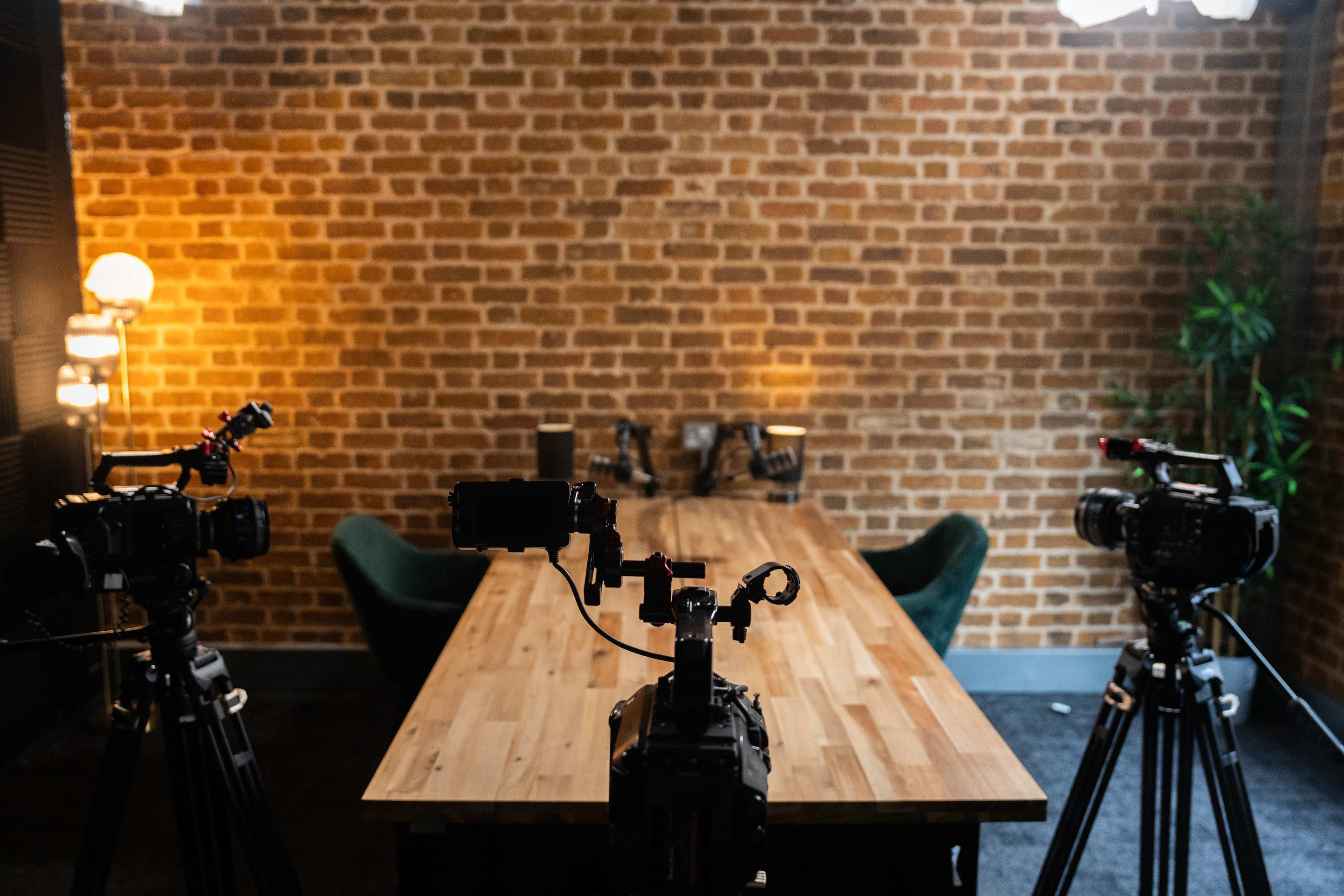 A well-lit, spacious room with a wooden table and three cameras positioned on tripods facing the table, against a brick wall backdrop.