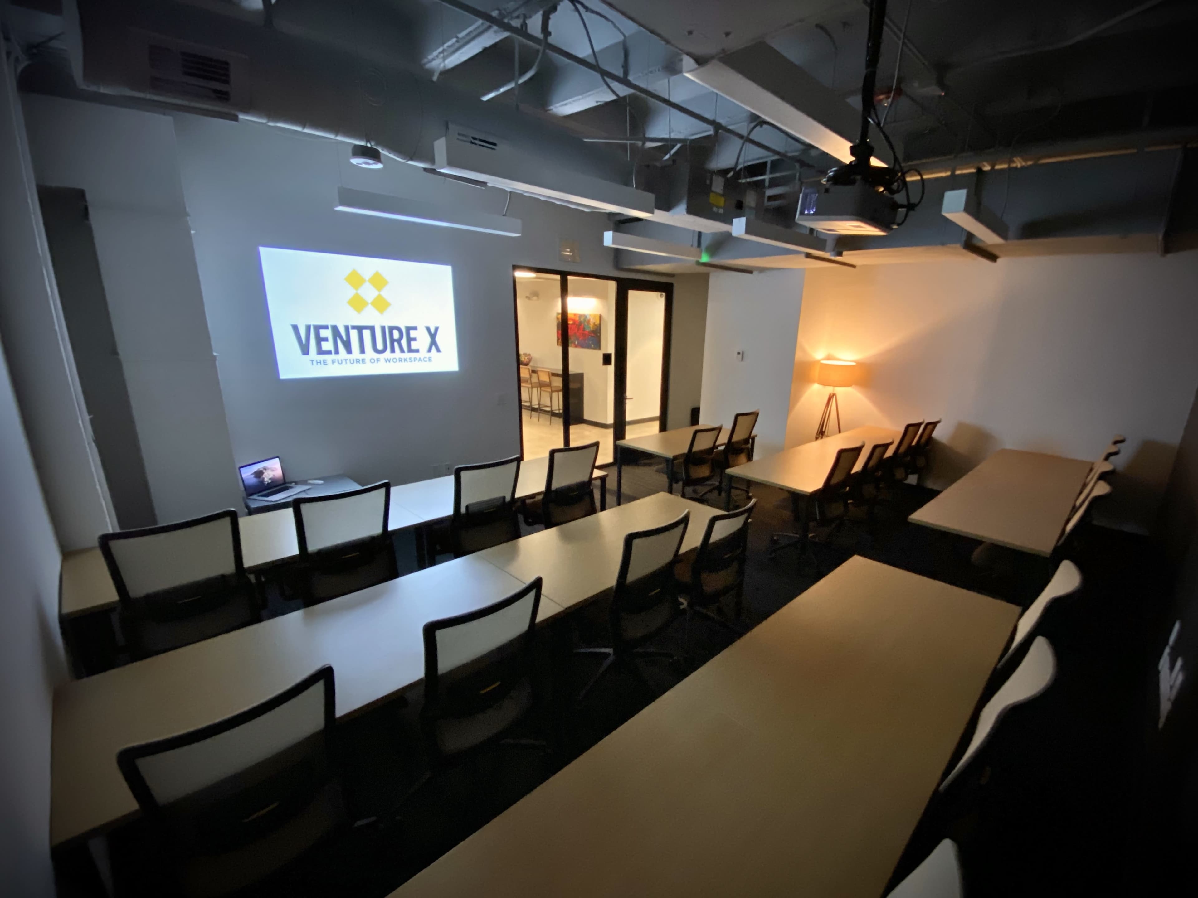 A conference room with rows of tables and chairs, featuring a projector displaying the "Venture X" logo on the wall.