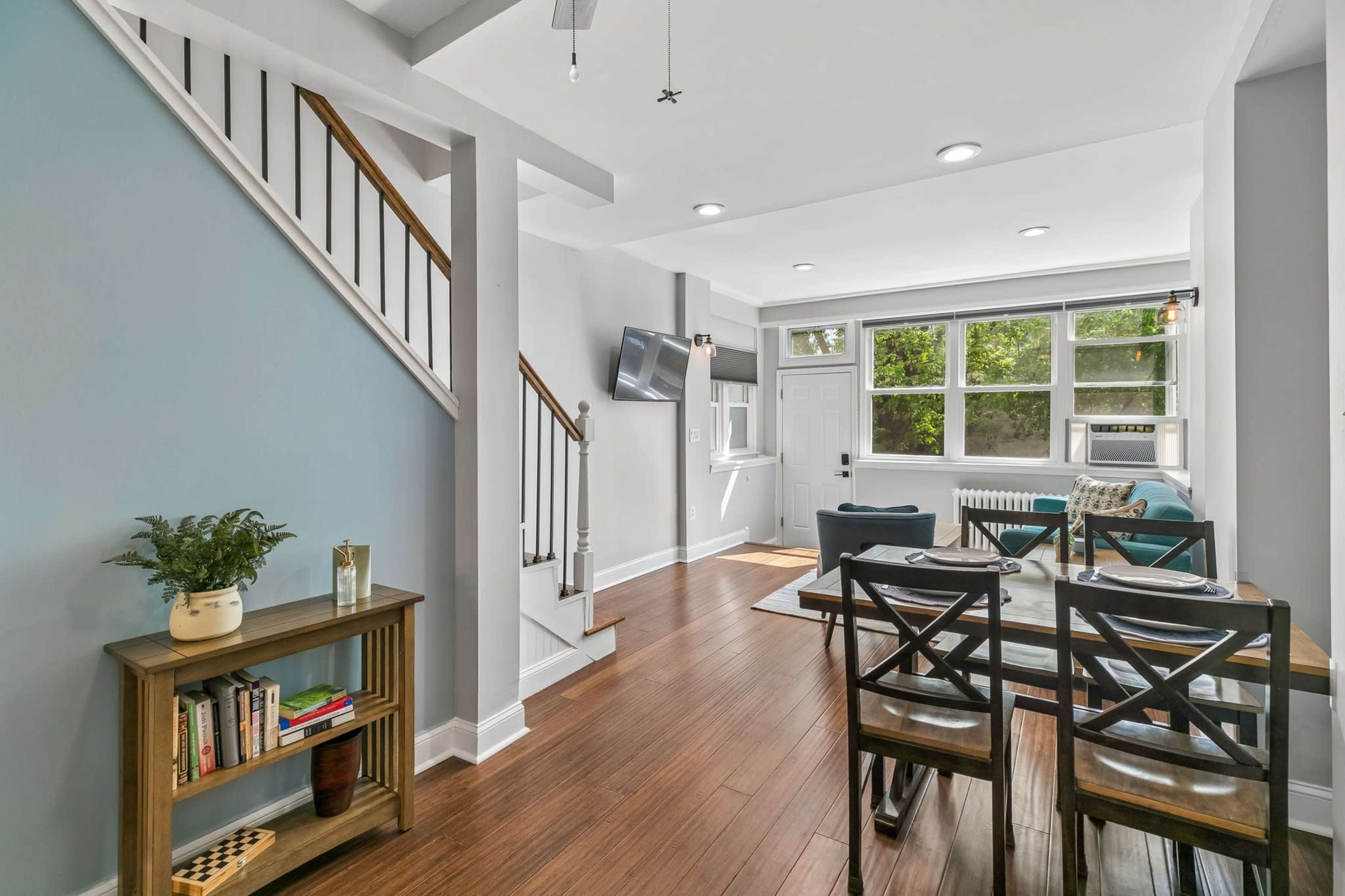 The image shows a modern interior of a living space featuring a dining area with a table and chairs, a staircase leading to an upper level, and large windows allowing natural light.