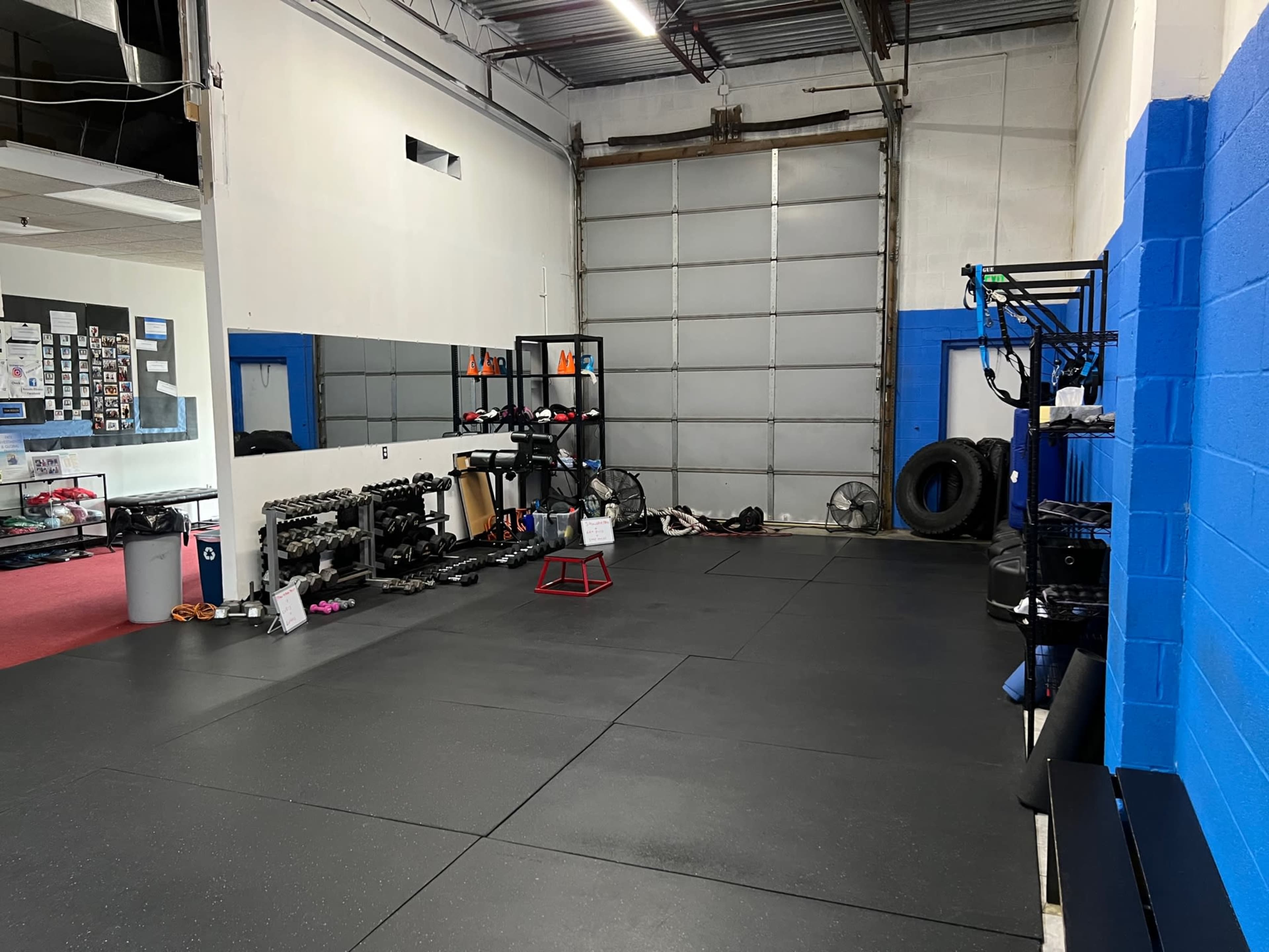 A fitness training area with rubber flooring, exercise equipment along the walls, and a garage door in the background.