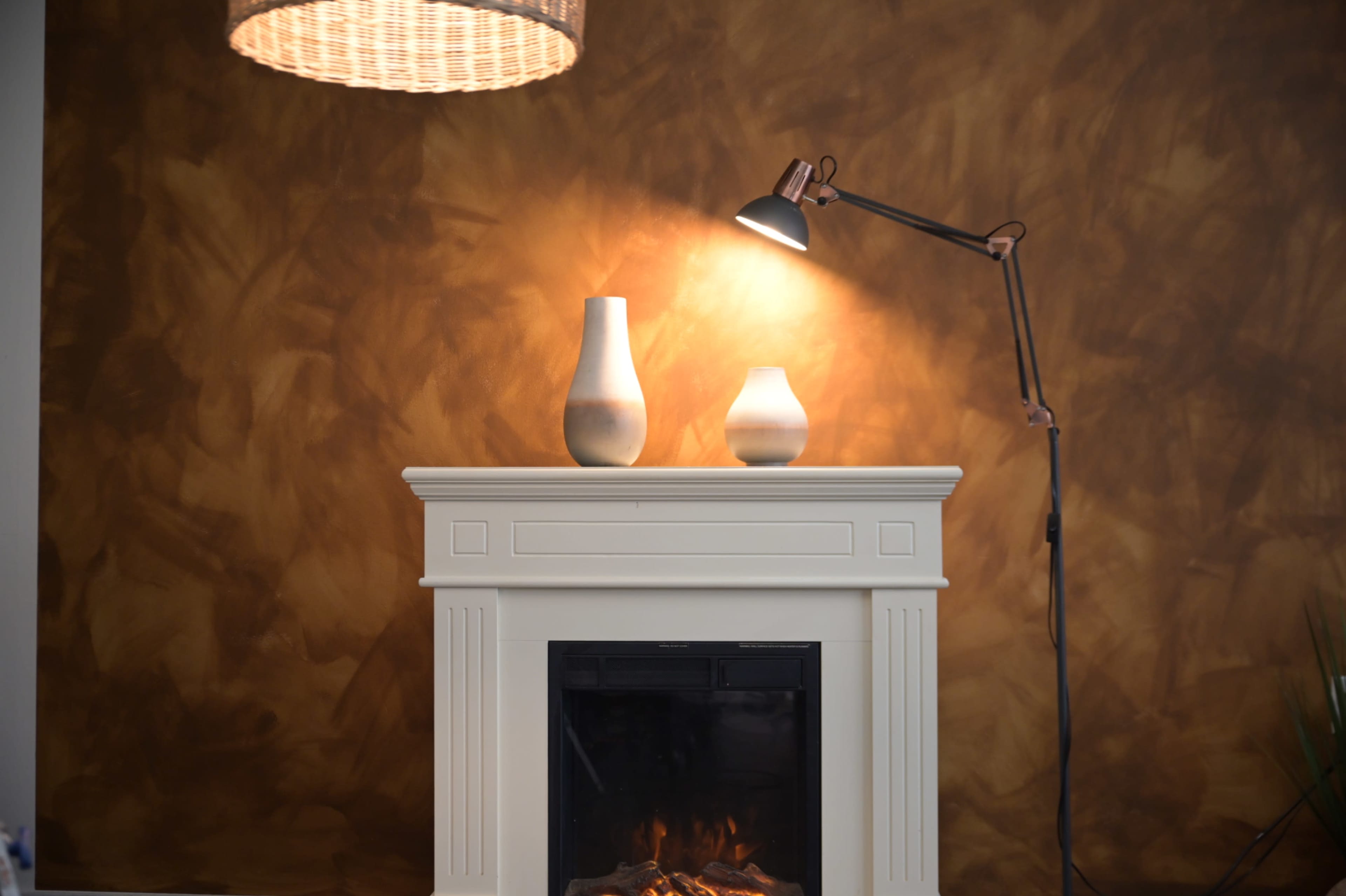 A white fireplace with two vases on top is illuminated by a lamp against a textured brown wall.