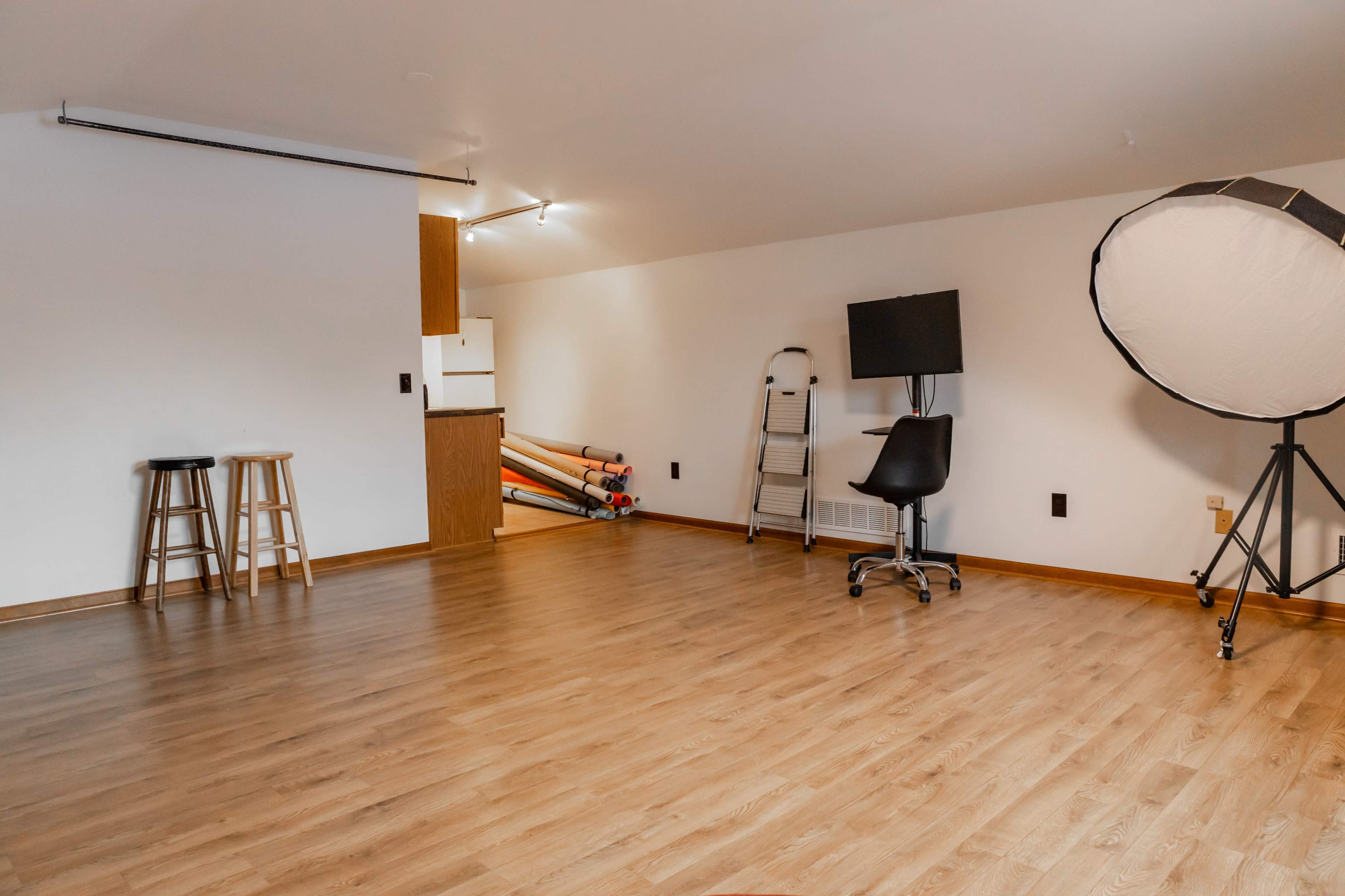 An empty room with wooden flooring, a kitchenette in the background, a tall standing light, a black chair on wheels, a folding ladder, and rolls of backdrop paper against the wall.