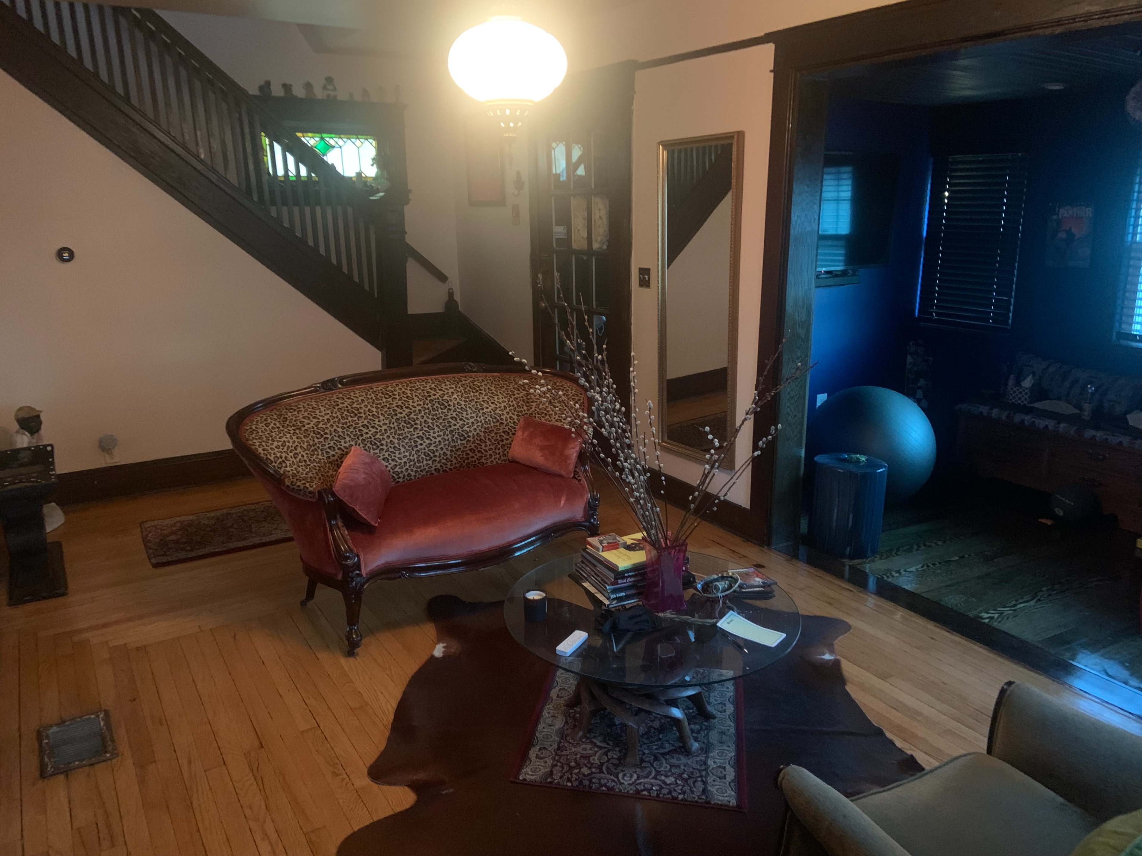 A furnished living room with a patterned sofa, a glass coffee table, and a staircase in the background, leading to a darkened area with blue walls.
