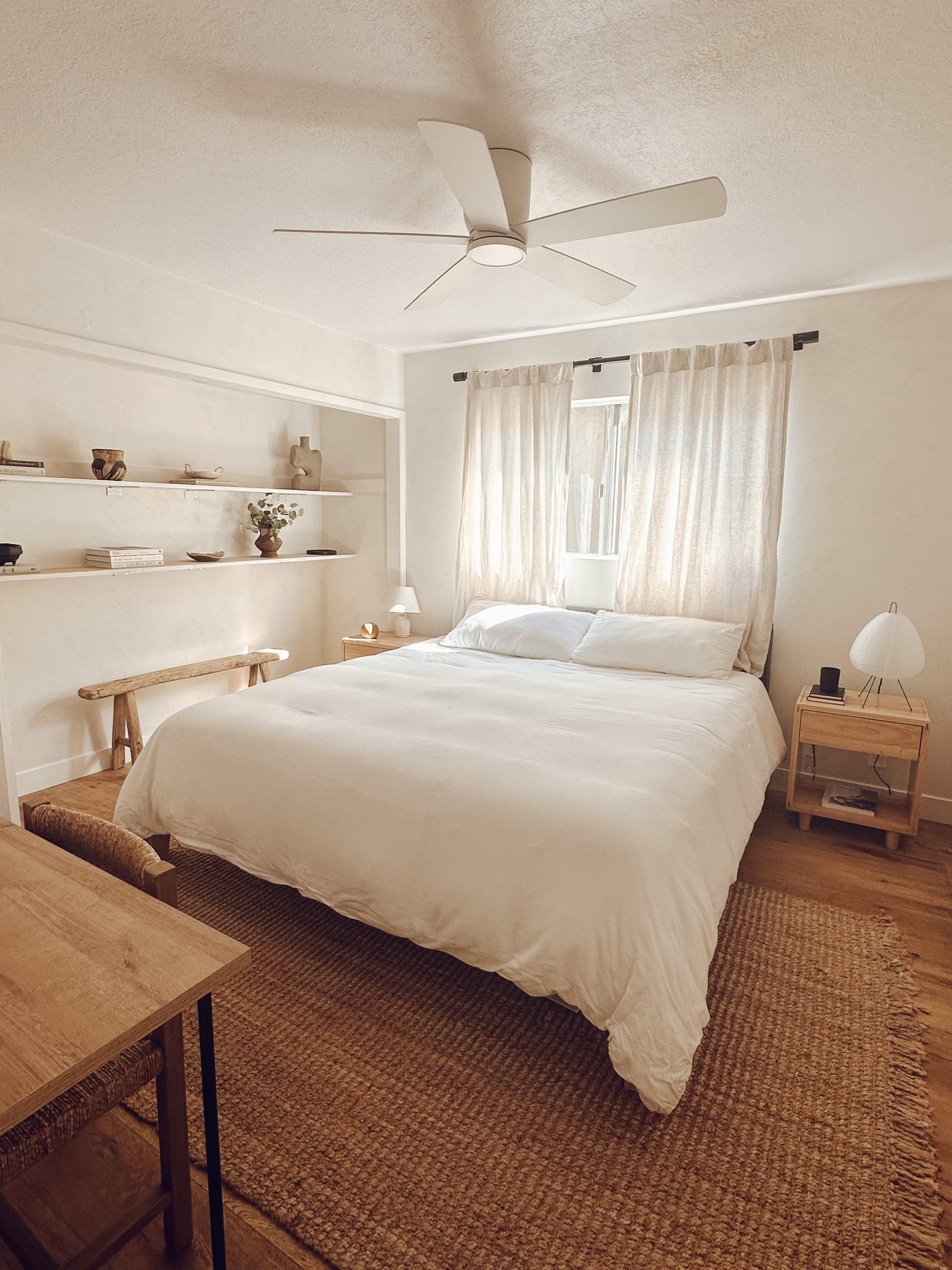 A simple bedroom with a large bed, a bedside table, a desk, and natural light streaming through sheer curtains.