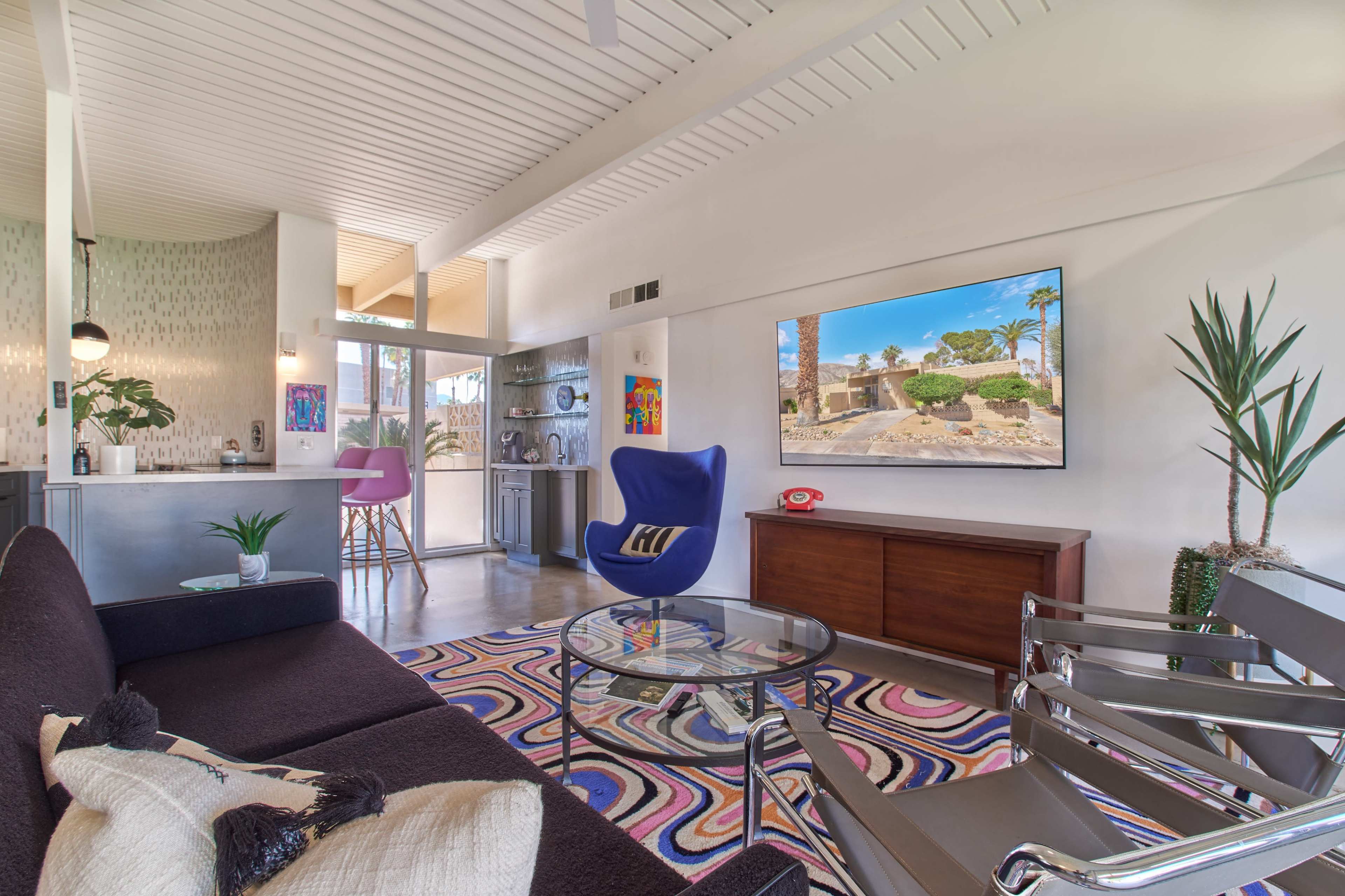 The modern living room features a black sofa, a glass coffee table, and a wall-mounted television displaying an outdoor scene, with a purple chair and decorative plants in the space.