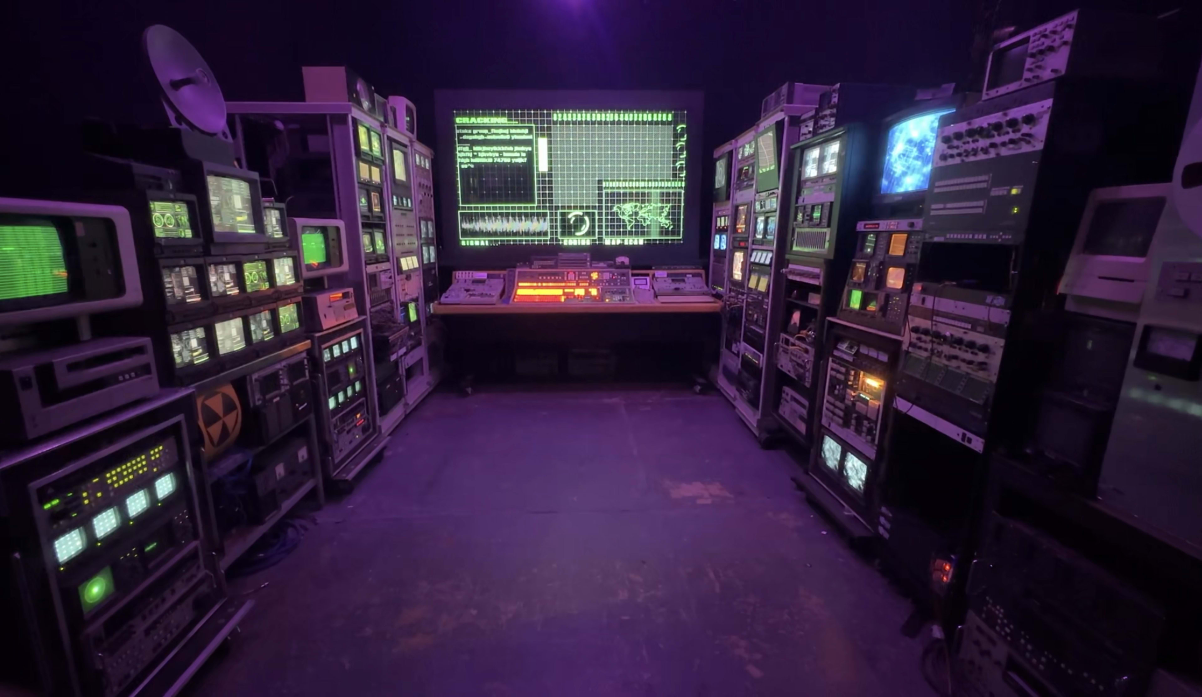 The image shows a dimly lit room filled with vintage computer equipment and screens, centered around a large monitor displaying green text and graphics.