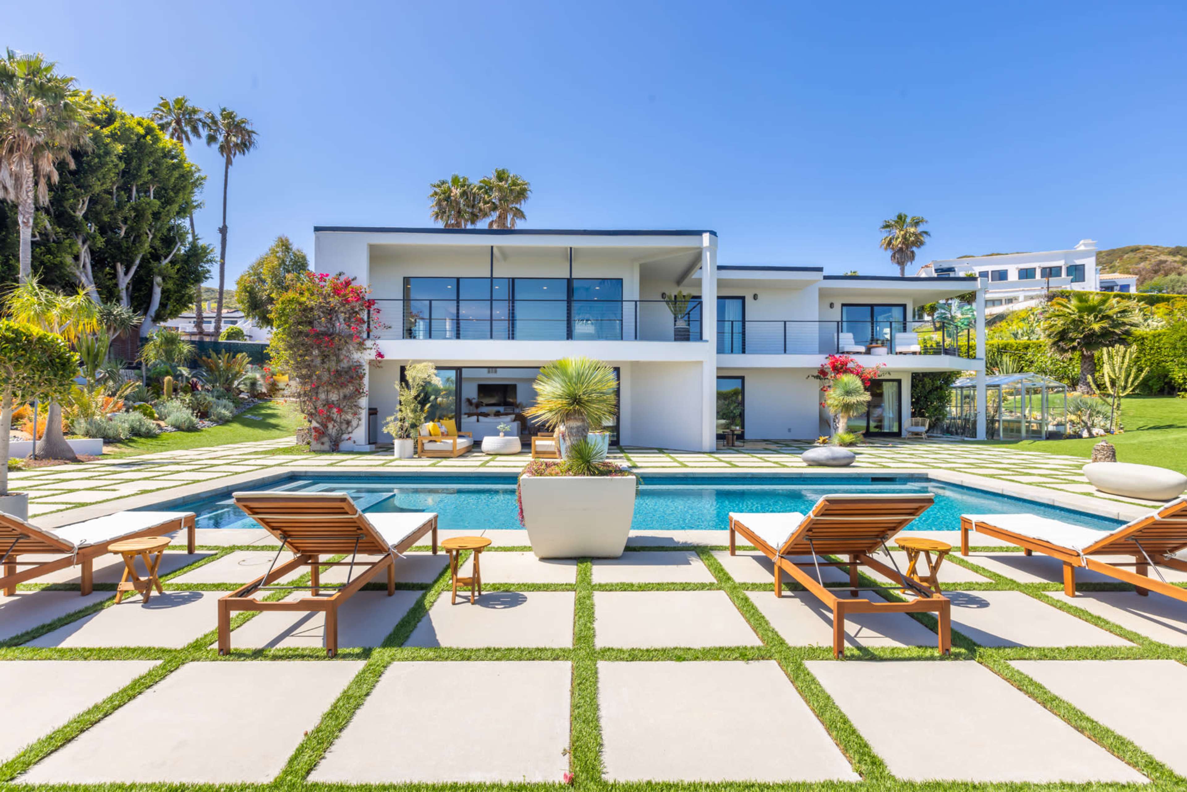 A modern two-story house with a swimming pool and lounge chairs is surrounded by landscaped gardens and palm trees.
