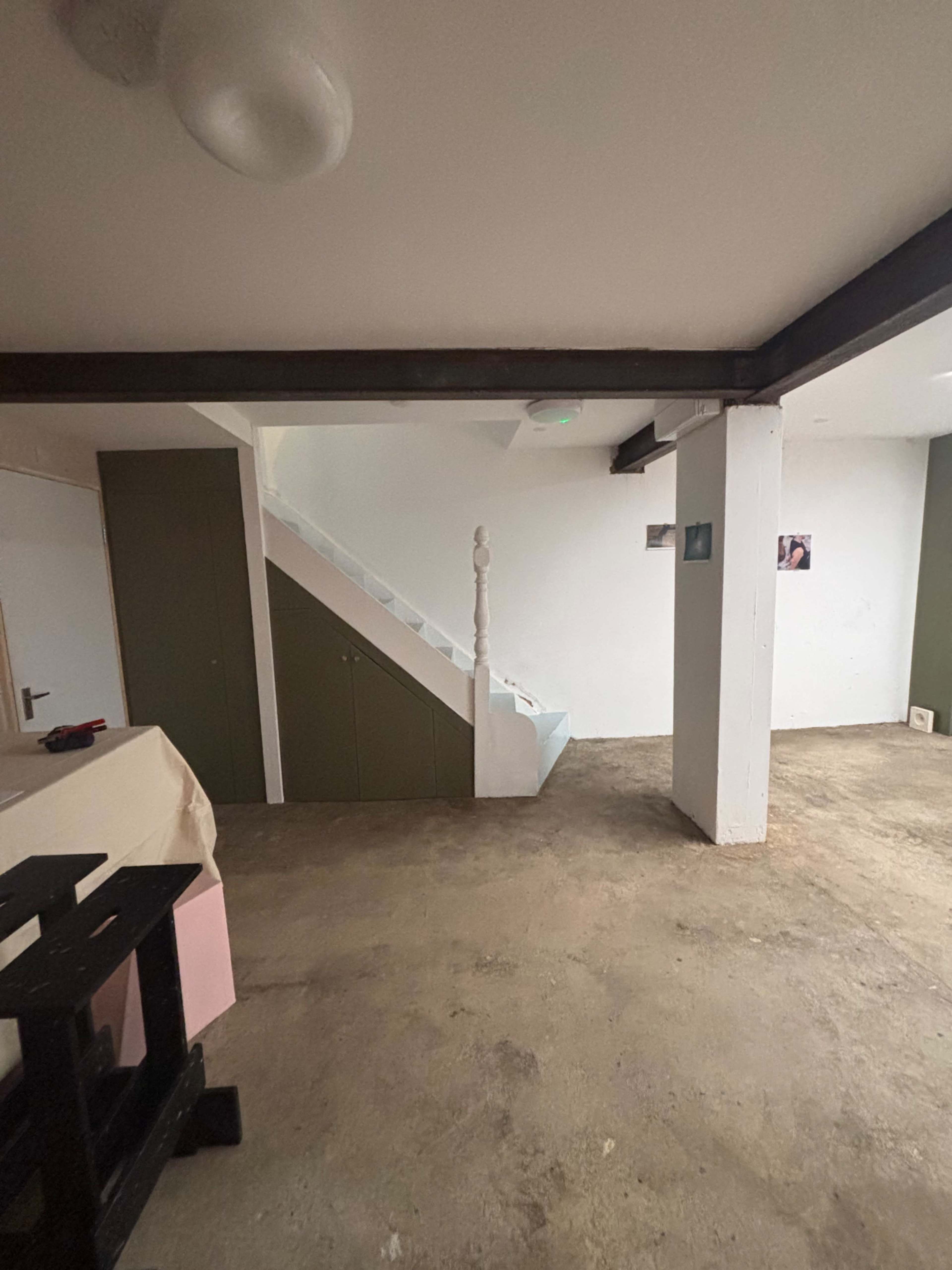 The image shows a sparsely furnished interior space featuring a staircase, white walls, and exposed beams.