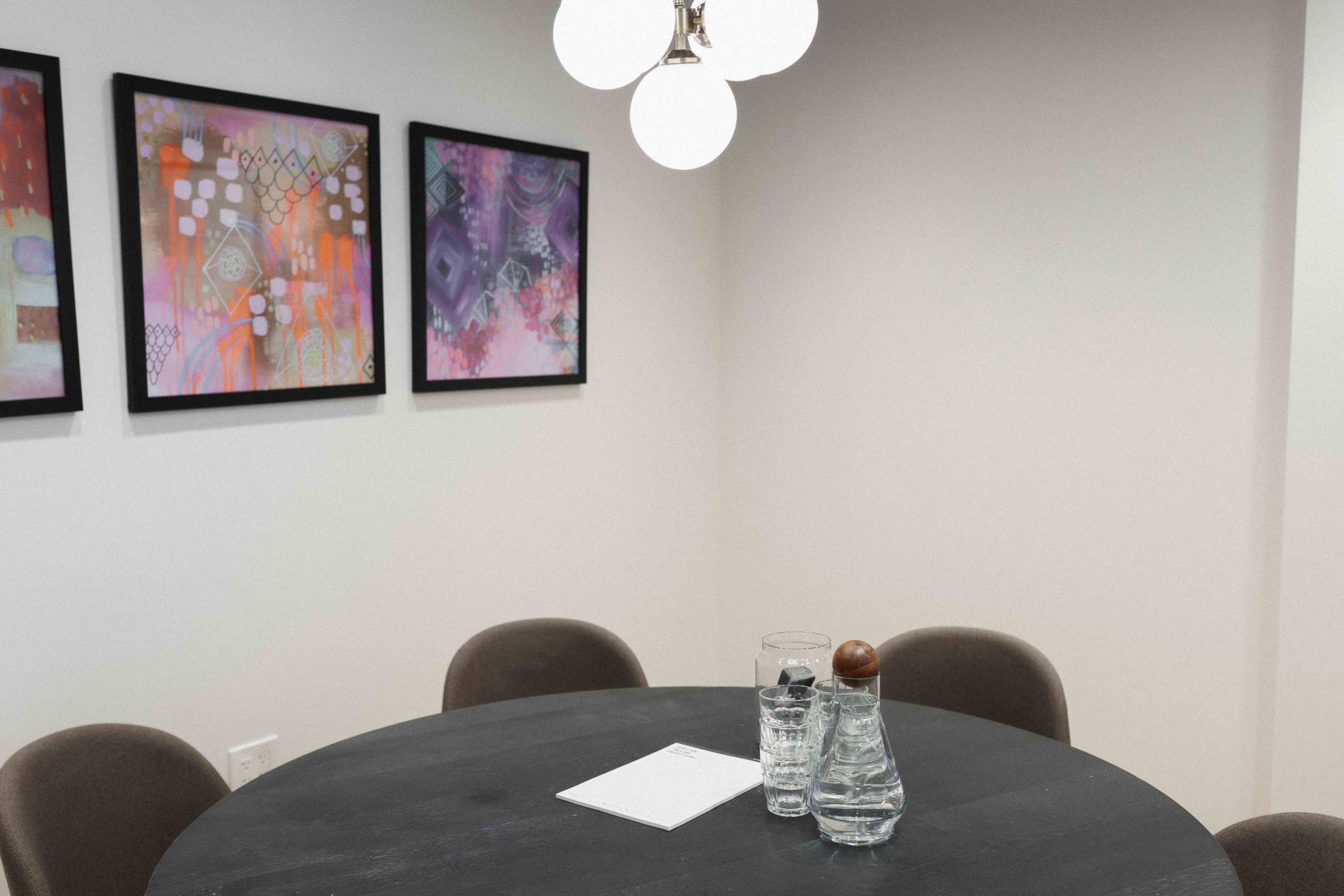A round black table is set in a room with four chairs and decorative artwork on the walls, featuring a water carafe and glasses on the table.
