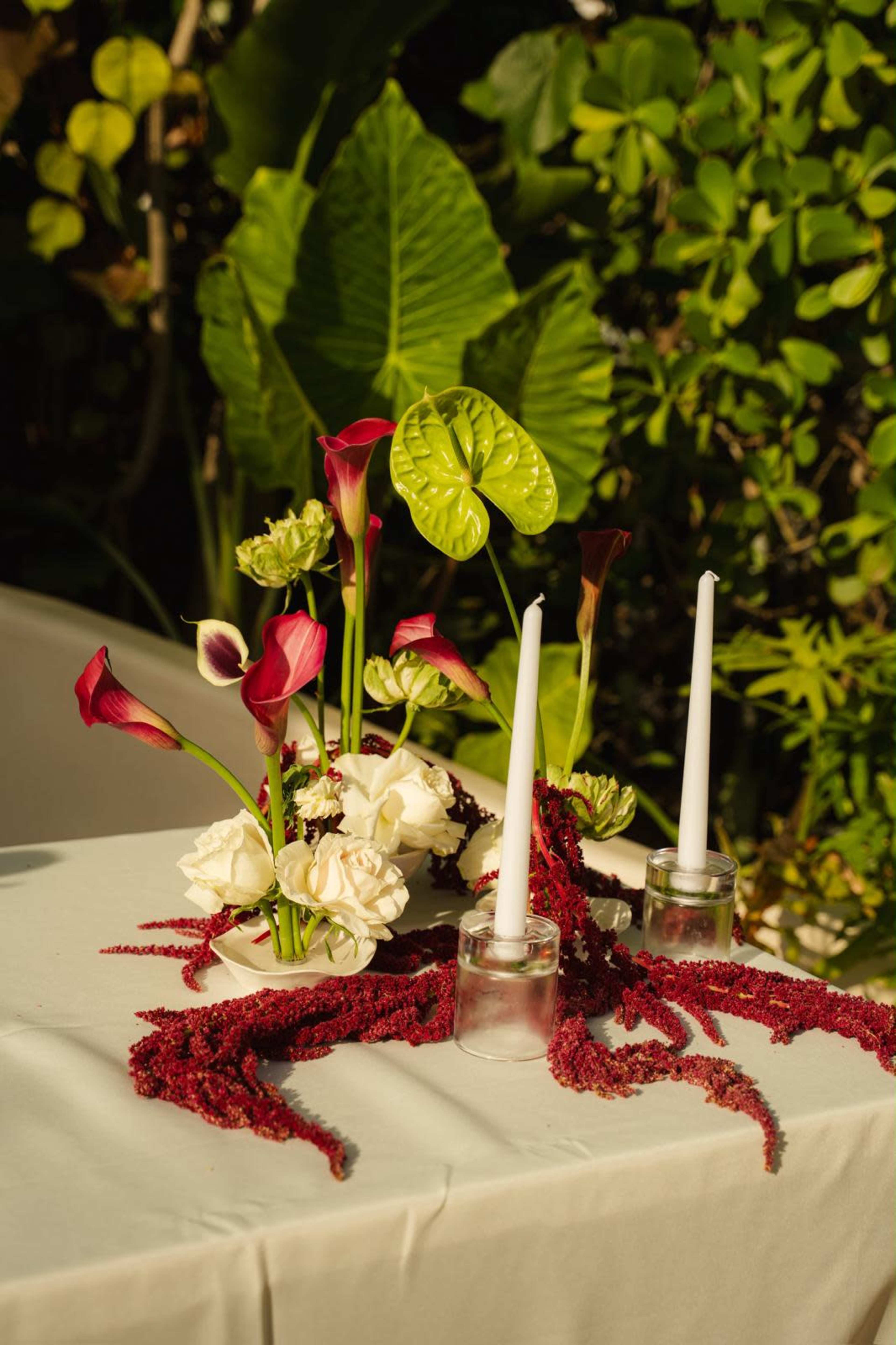 A table centerpiece features a floral arrangement with red and white flowers, complemented by two white candles and a decorative red garland, set against a lush green backdrop.