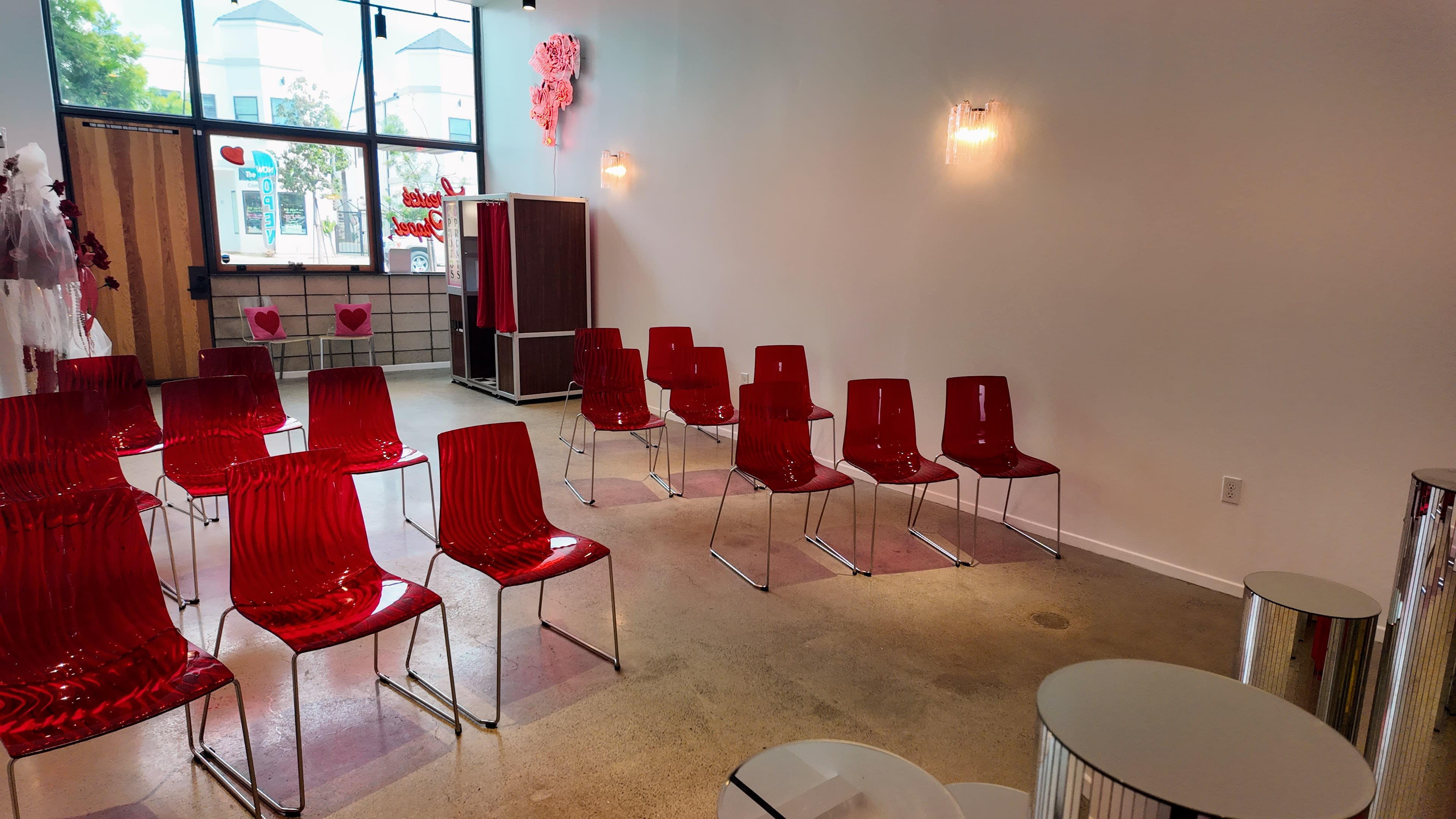A room with a row of red plastic chairs arranged in front of a blank wall and small circular tables nearby.