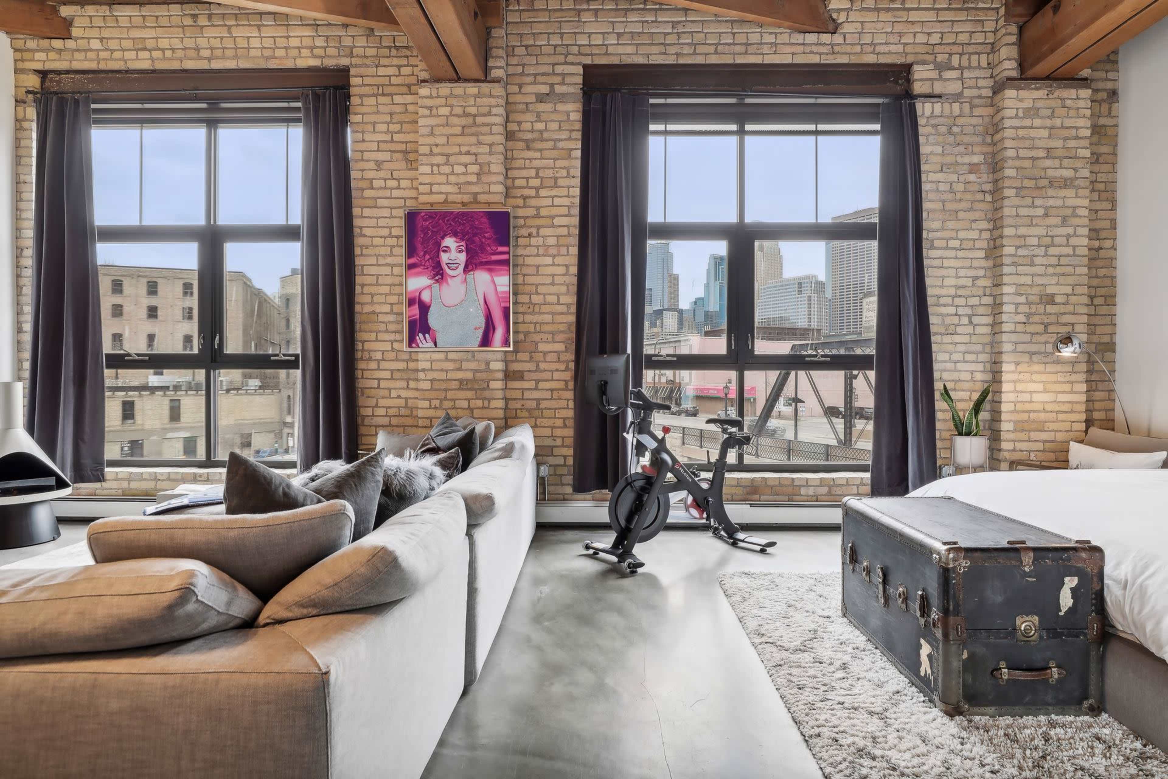 A spacious loft with exposed brick walls features large windows, a workout bike, a vintage trunk, and a sofa in front of a bed.