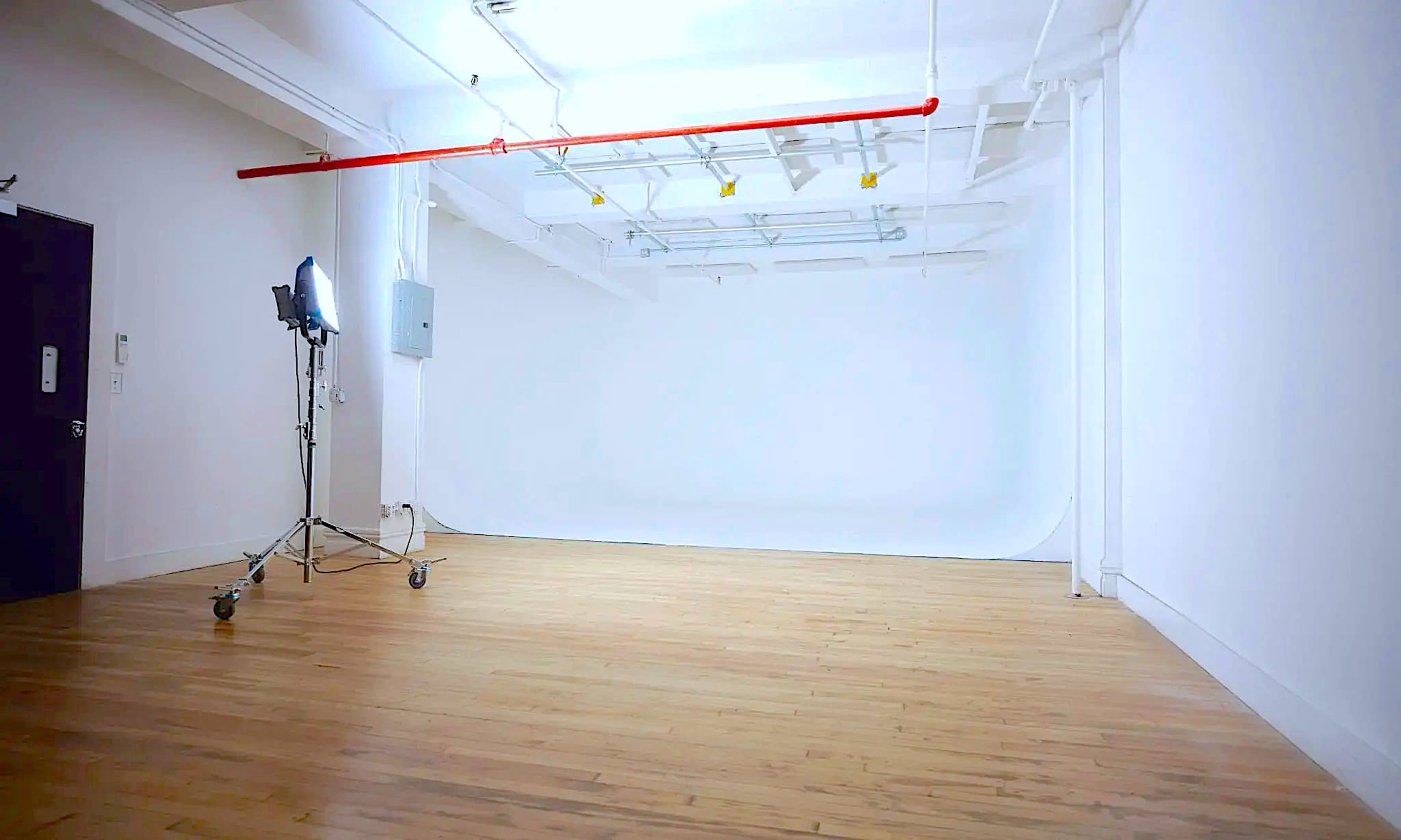 The image shows a spacious, empty studio with wooden floors and a white backdrop, illuminated by a softbox light on wheels.