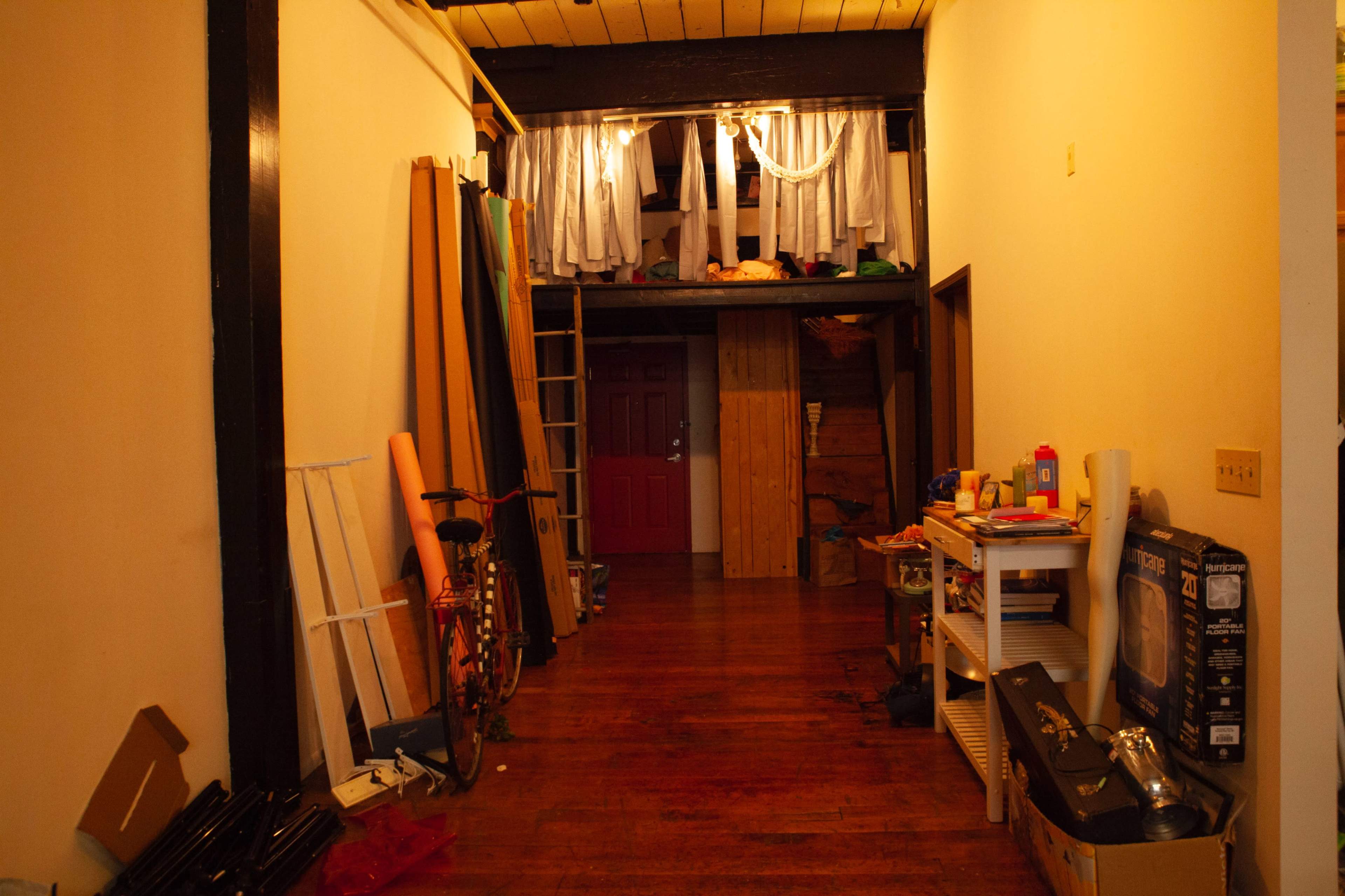 The image shows a narrow hallway lined with various materials and items, leading to a red door at the far end and a lofted area above with draped fabric.
