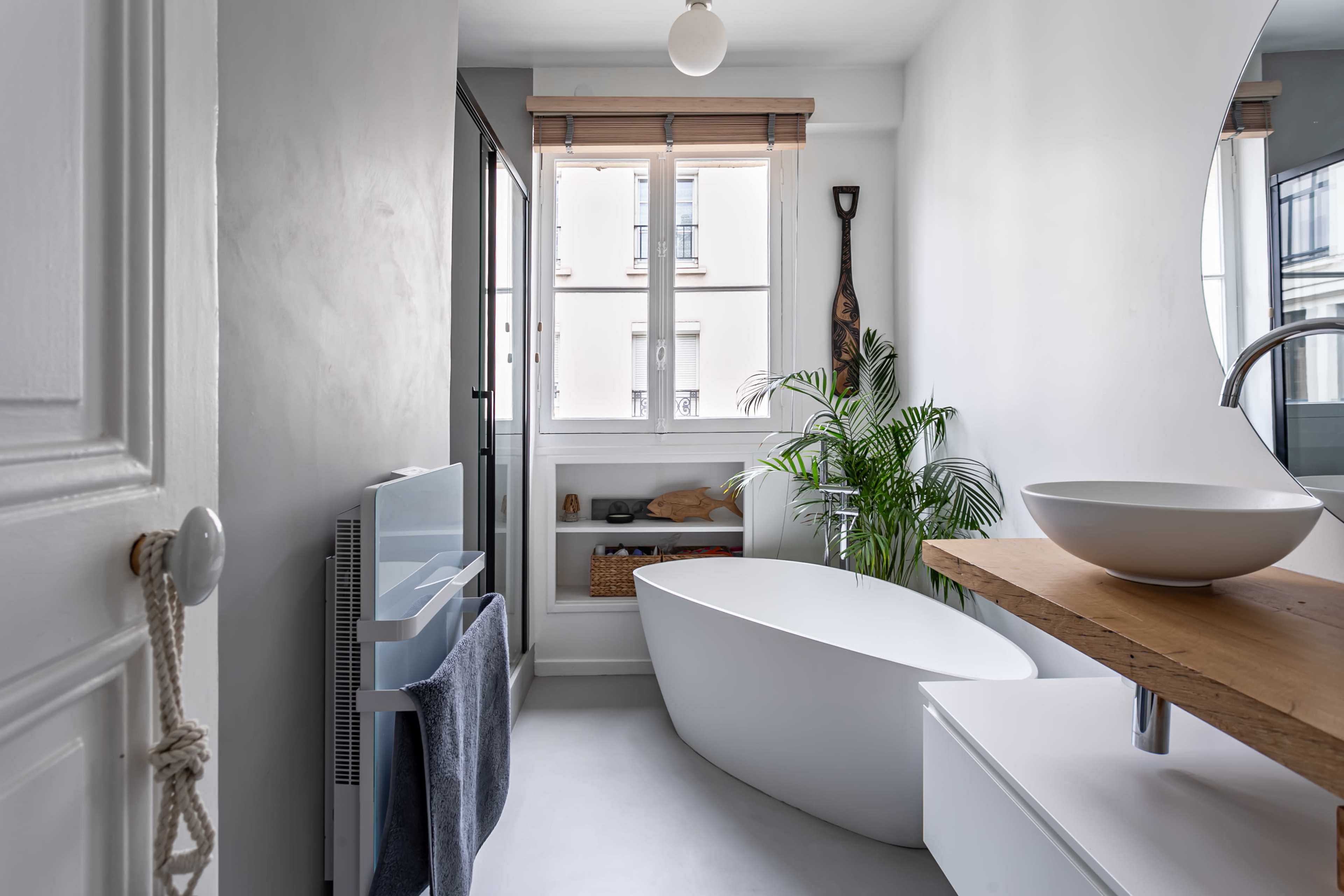 A modern bathroom features a freestanding tub, a wooden counter with a bowl sink, and large windows allowing natural light, complemented by indoor plants.