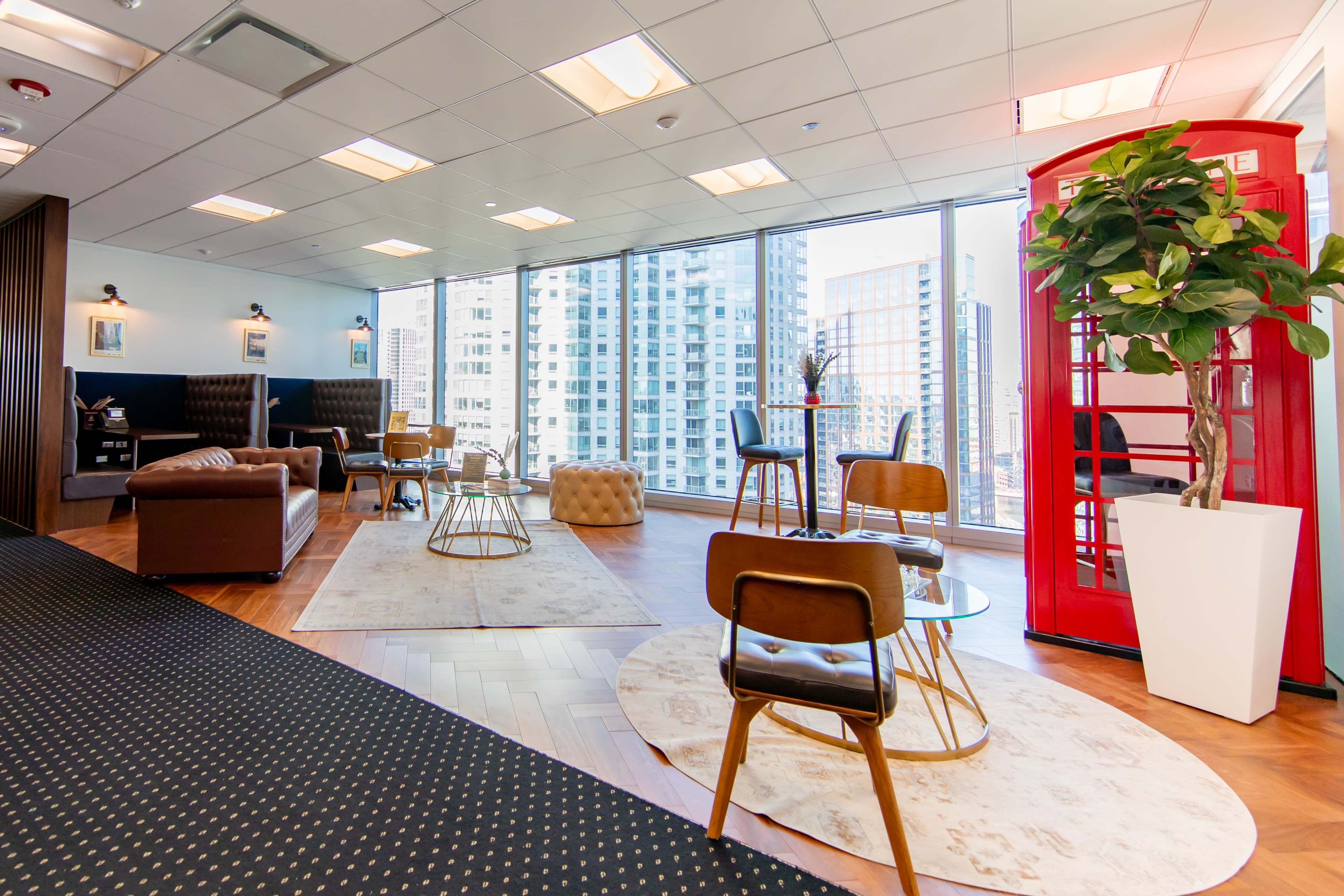 The image shows a modern office lounge with large windows, featuring a red telephone booth, comfortable seating, and stylish decor.