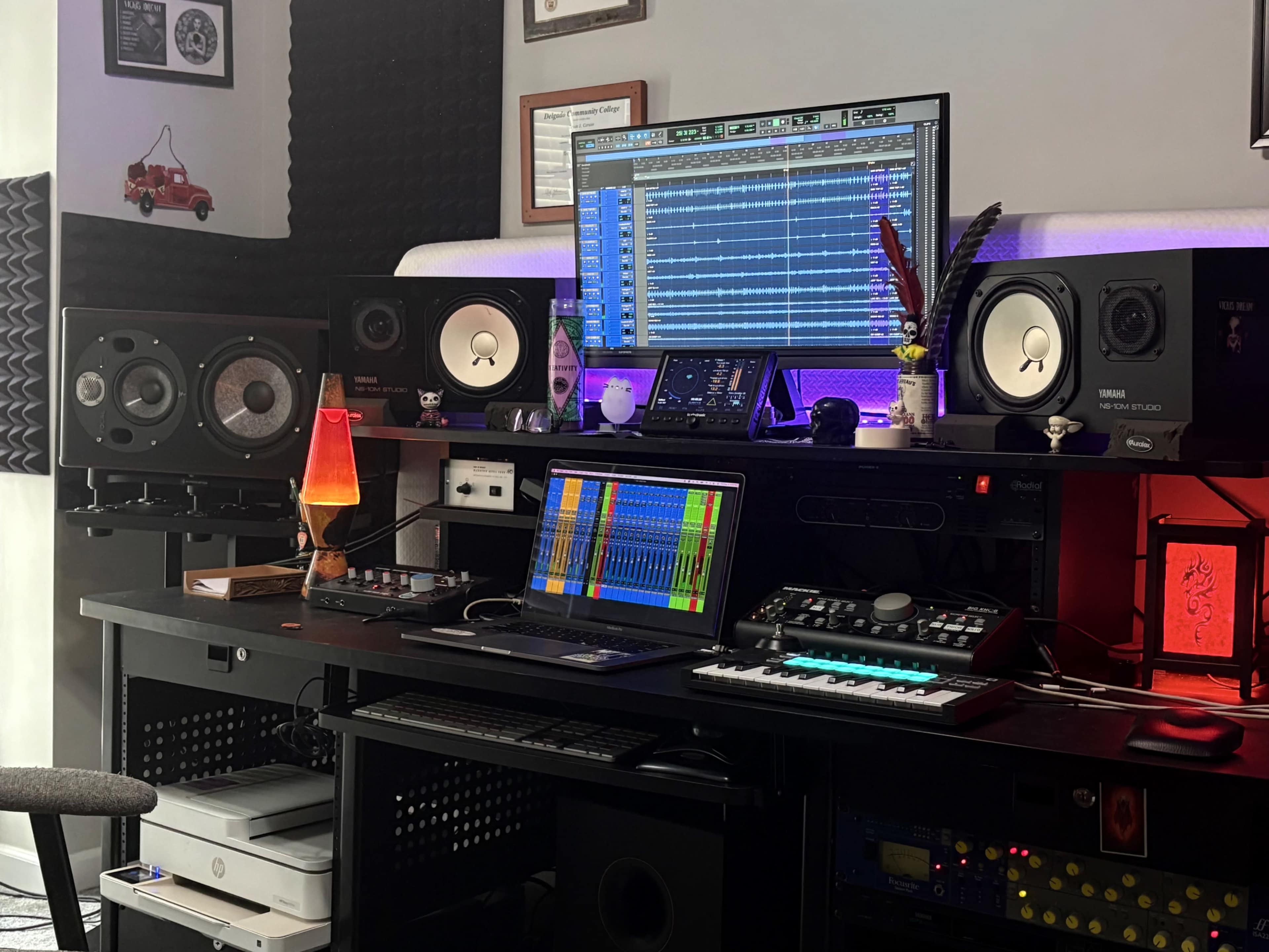 The image shows a home music studio setup featuring a computer monitor displaying audio software, speakers, a MIDI keyboard, and various audio equipment on a dark workstation.