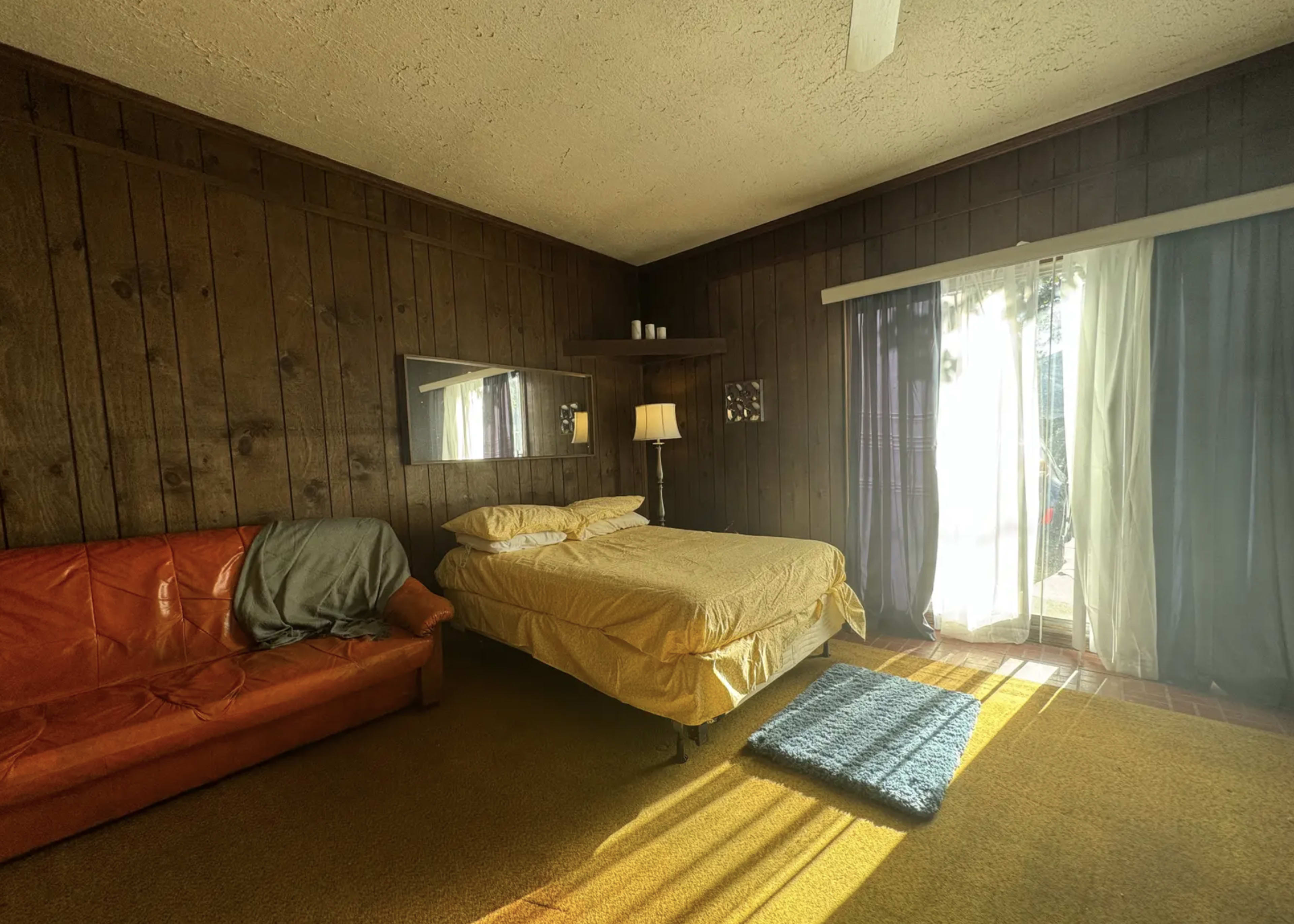 The image shows a cozy bedroom with wood-paneled walls, a bed with yellow bedding, a brown couch, and large windows covered by sheer curtains allowing sunlight to filter in.