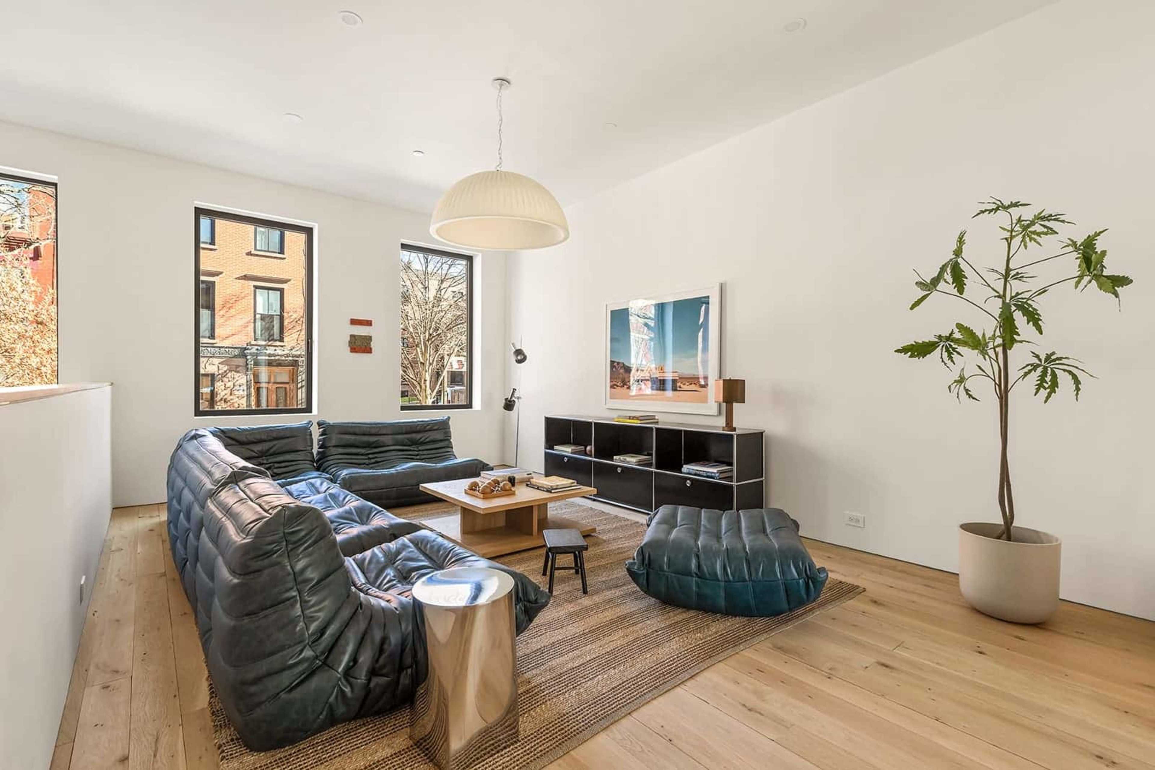 The image shows a modern living room with a sectional sofa, a coffee table, and a media console against a wall with large windows providing natural light.
