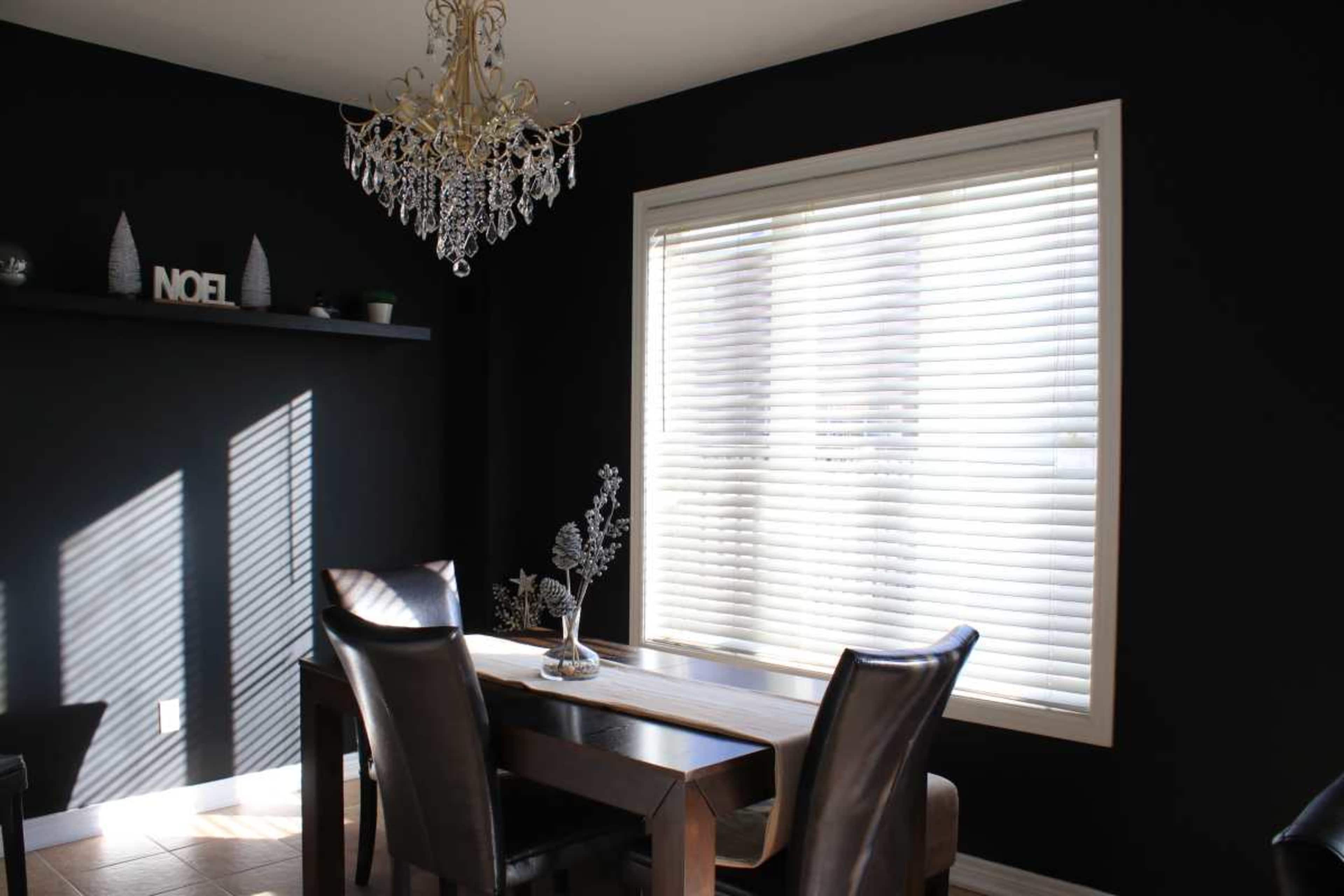 A dining room features a wooden table, black leather chairs, and a large window with blinds that allows light to enter, accompanied by a chandelier overhead.