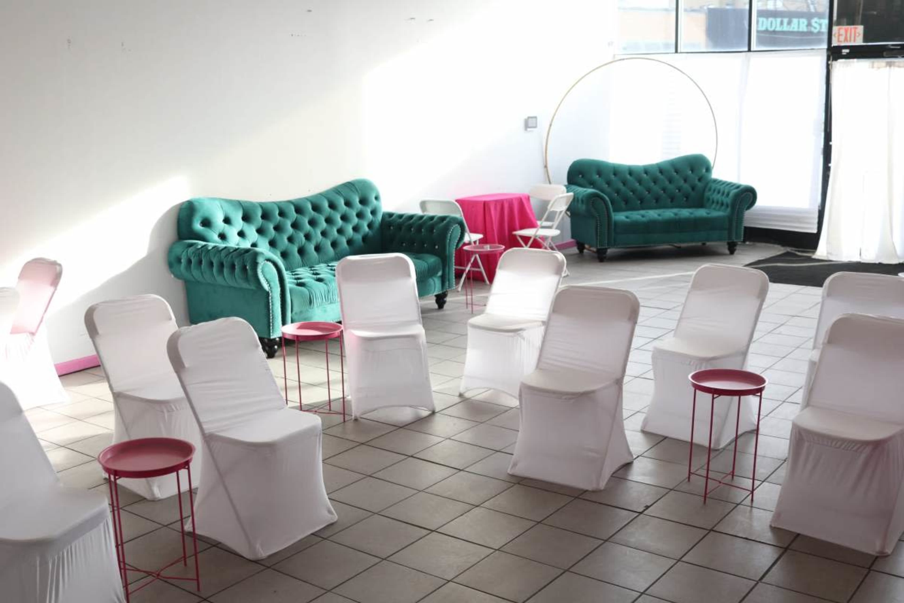 The image shows a brightly lit room with two teal tufted sofas, several white-covered chairs arranged in a circle, and small pink side tables.
