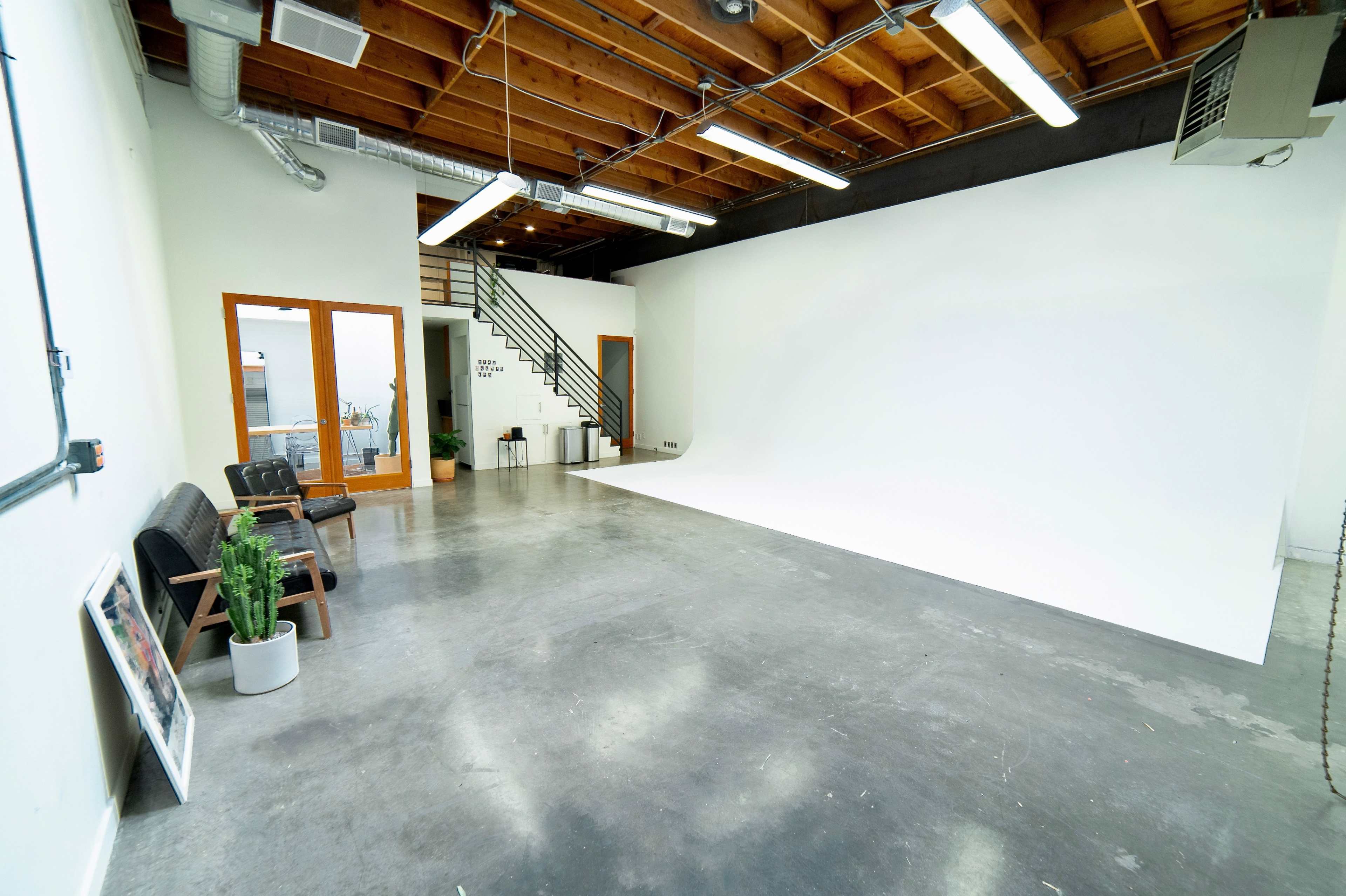 A spacious studio with a white backdrop, concrete flooring, and wooden ceiling beams, featuring a staircase and glass doors leading to another room.